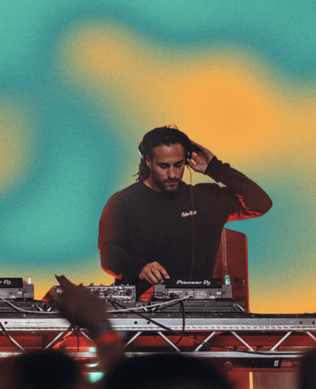 A DJ in a black shirt stands behind a mixing desk, one hand on the headphones and the other adjusting controls, with a vibrant, blurred yellow and teal background.