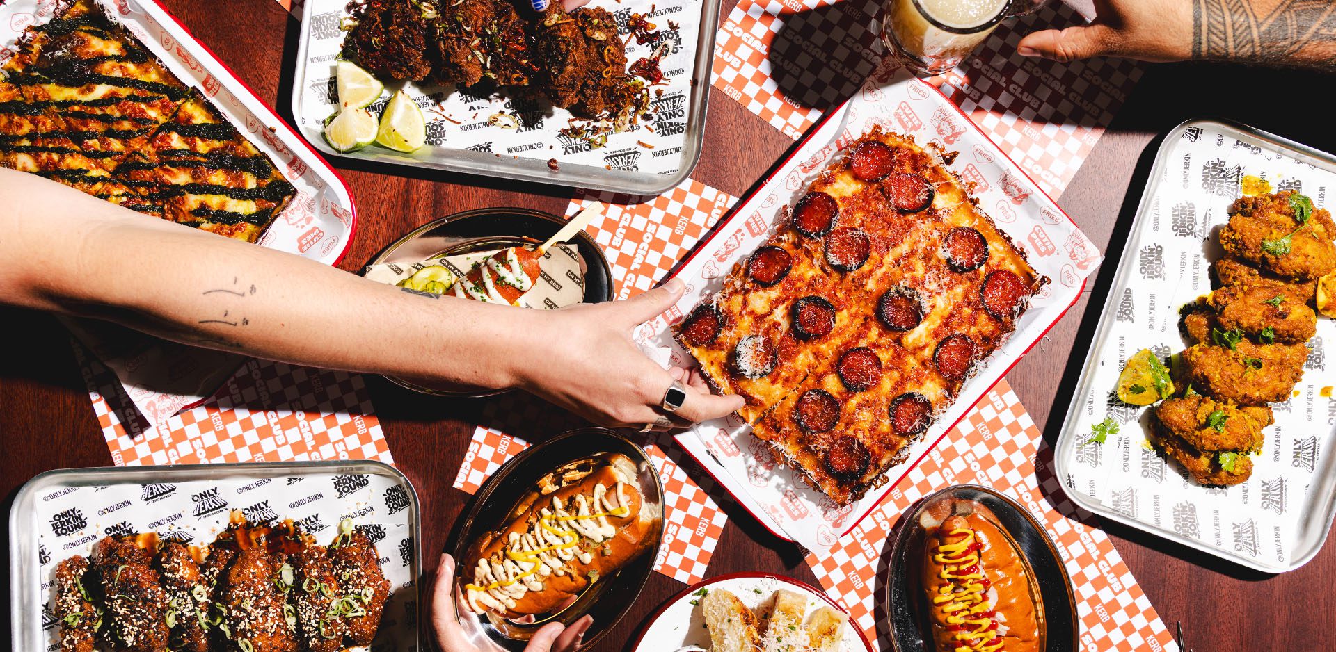 A festive table covered with Christmas favourites, including Detroit-style pepperoni pizza, sauced chicken wings, grilled sweetcorn, fried gherkins, a hot dog, and hands reaching for food on patterned paper trays.