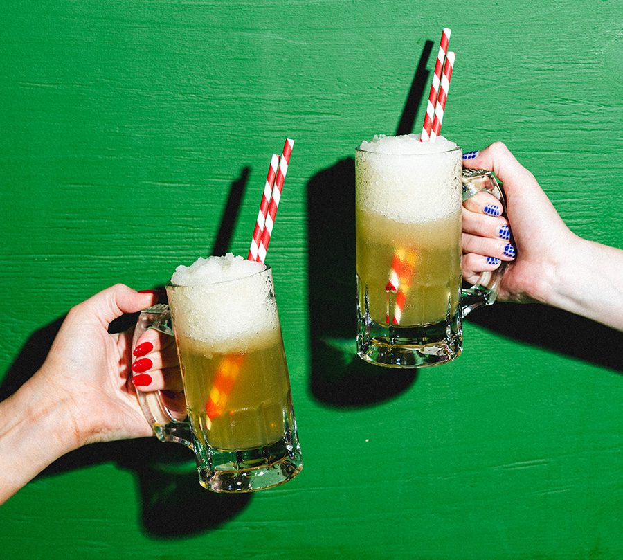 Two hands with colourful manicured nails hold frothy mugs of a yellow drink with red and white striped straws, raised in a toast against a bright green background.