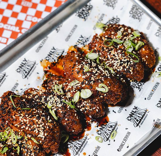 A tray of crispy fried chicken wings coated in sauce, topped with sesame seeds and sliced spring onions, served on branded paper at Food Kerb Social Club.