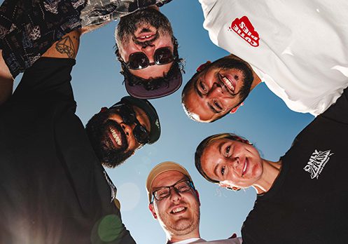 Five people stand in a circle, smiling and looking down at the camera against a clear blue sky. They appear happy and relaxed, dressed casually, with sunlight illuminating their faces.