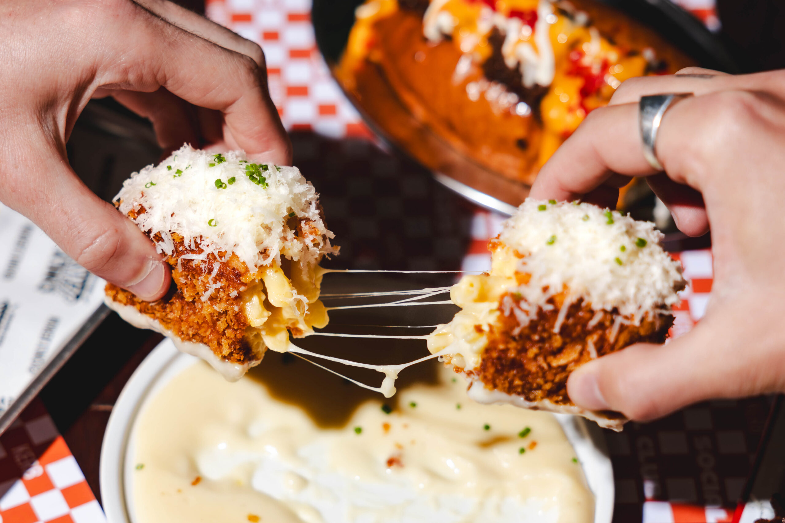 Two hands pull apart a fried, cheese-covered macaroni ball, with melted cheese stretching between the halves above a bowl of cheese sauce; another cheesy dish is blurred in the background.
