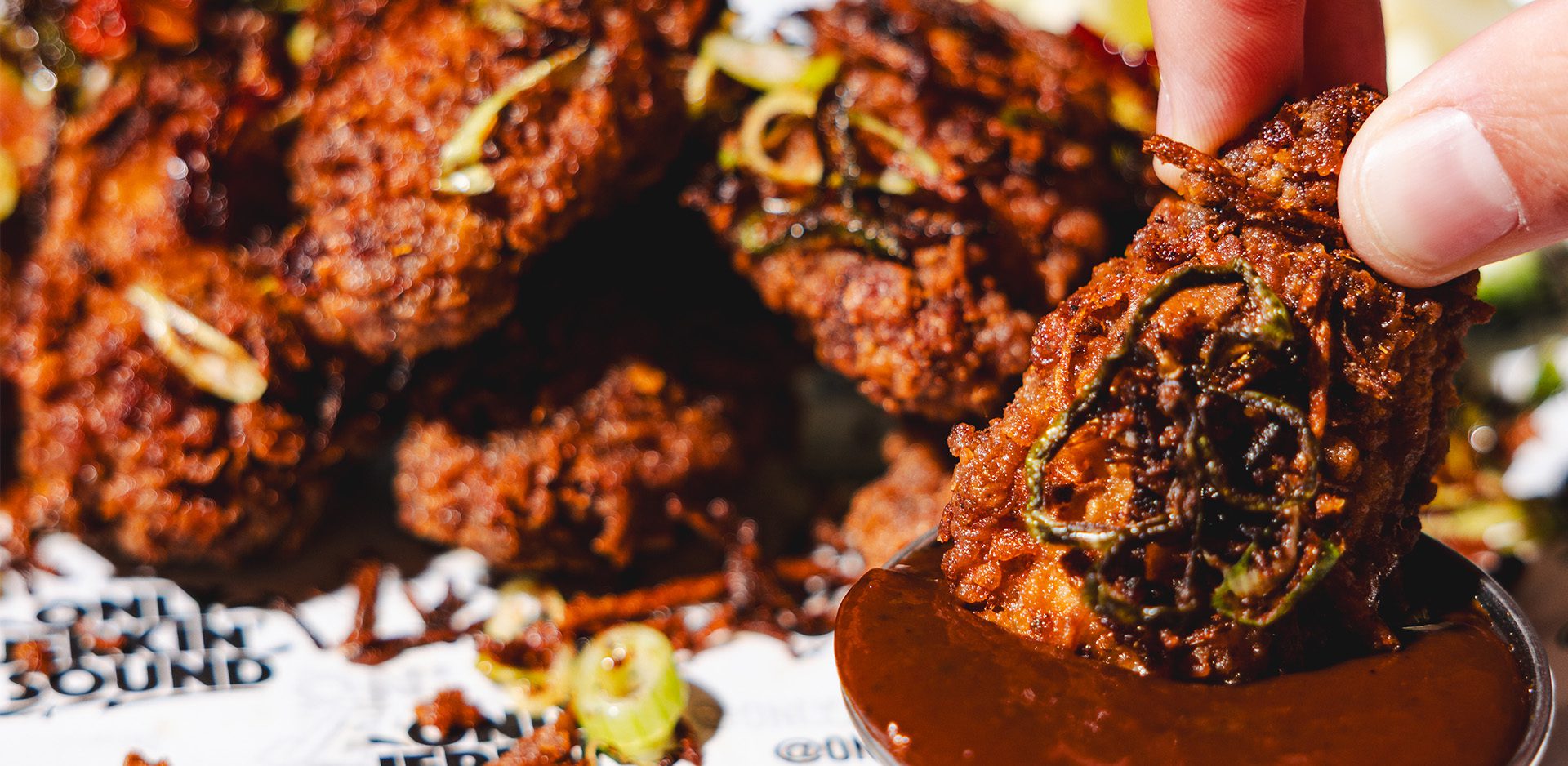 A hand dips a crispy, spicy fried chicken wing topped with sliced spring onions into a cup of brown sauce at Food Kerb Social Club, with more wings piled in the background.