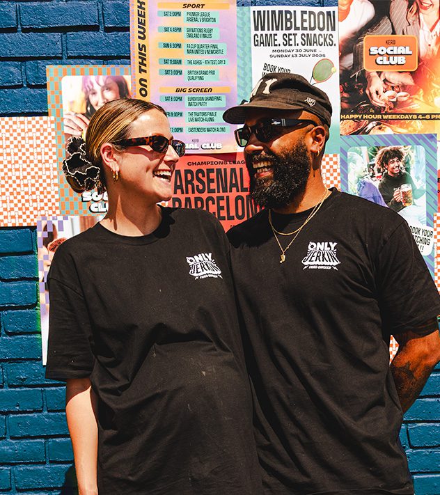 Two people wearing black Only Jerk shirts and sunglasses stand smiling at each other in front of a blue brick wall, covered with colourful event posters for Food Kerb Social Club.