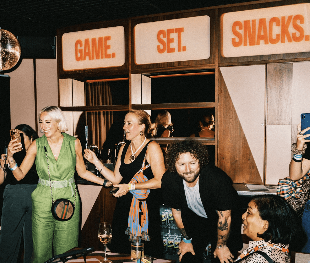 A group of people gathers at a party, laughing and taking photos in front of a retro sign that reads GAME. SET. SNACKS. Drinks and snacks are on the table, creating a festive atmosphere.