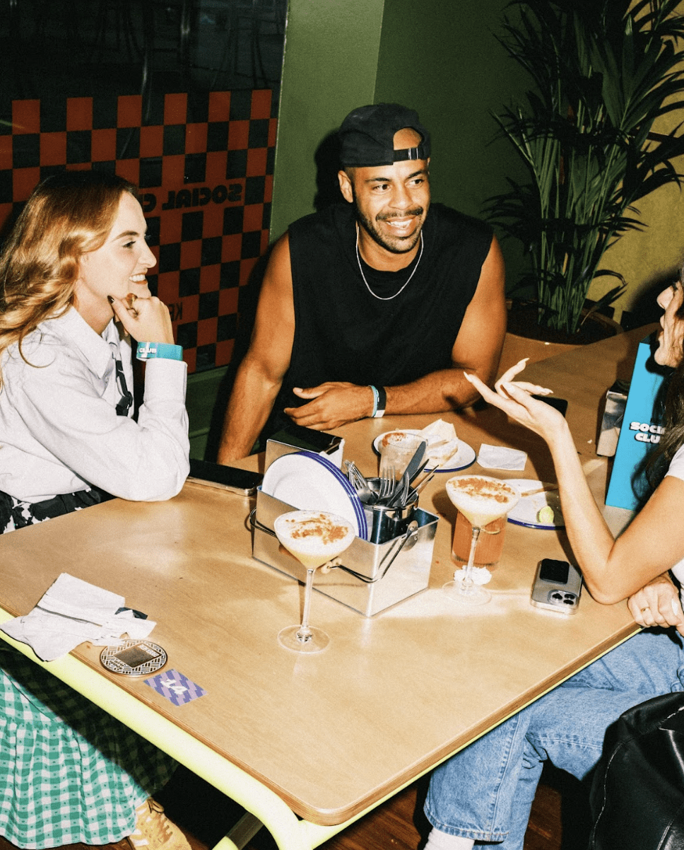 Three friends sit at a table in a restaurant, laughing and talking over cocktails. Cutlery and serviettes rest on the table, with plants and a chequered wall behind them—perfect moments when you book KERB Social Club.