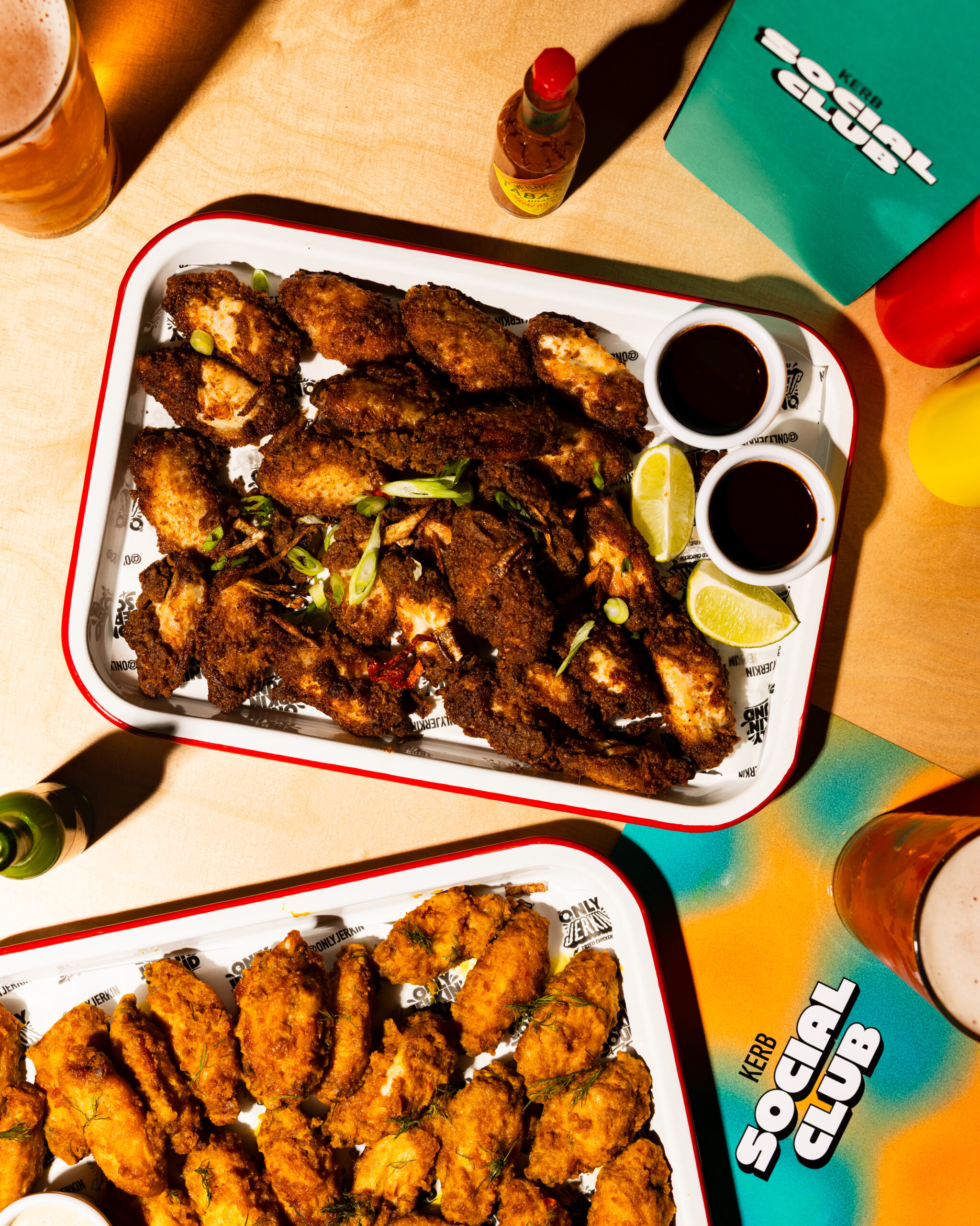A tray of crispy fried chicken wings garnished with spring onions, lime wedges, and dipping sauces sits on a table with drinks, hot sauce, and a green Social Club menu at this lively Sports Bar in Shoreditch. Another tray of wings is partially visible.