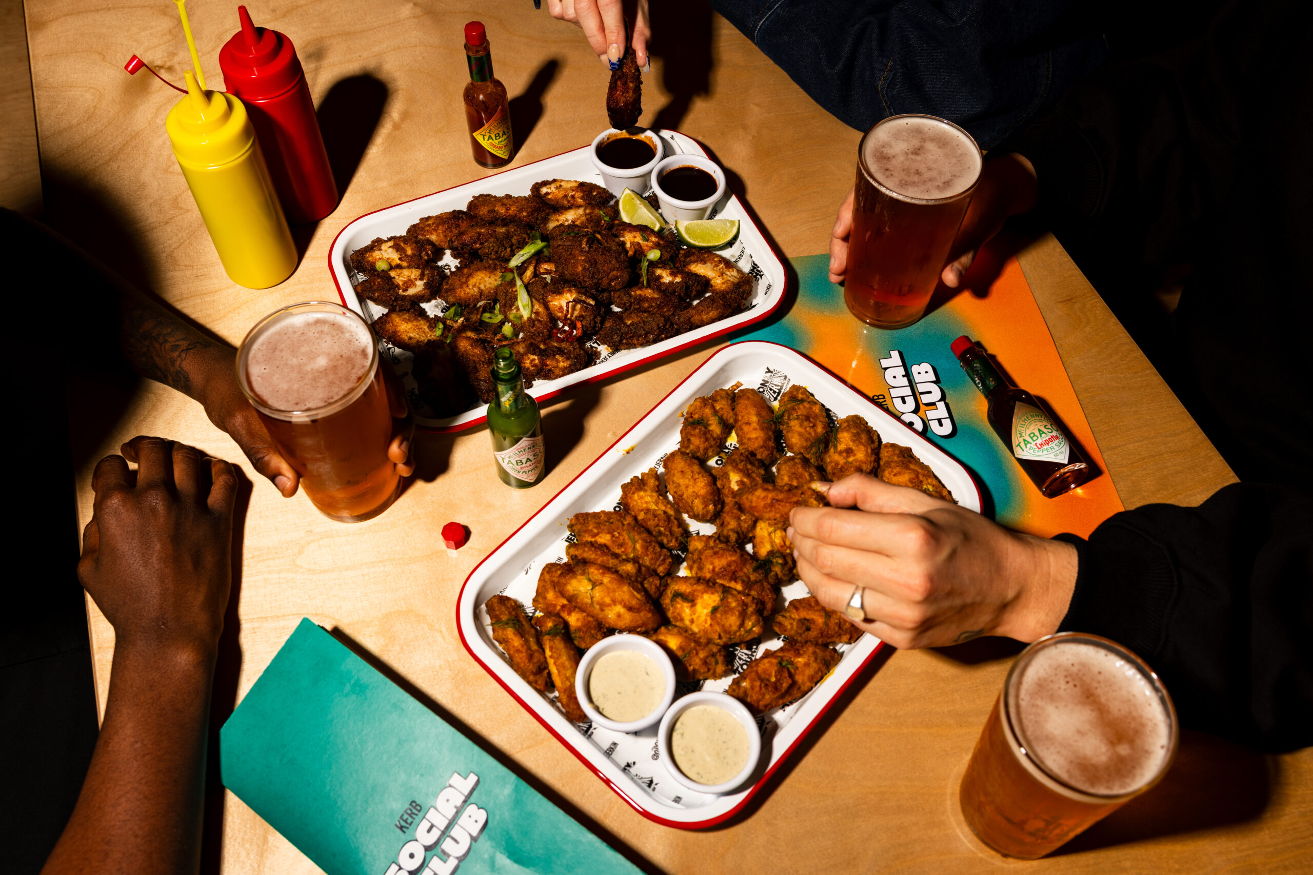 At a lively Sports Bar in Shoreditch, three people share trays of chicken wings, dipping sauces, and drinks on a wooden table with menus, hot sauce, and condiments. Hands reach for wings and hold glasses of beer.