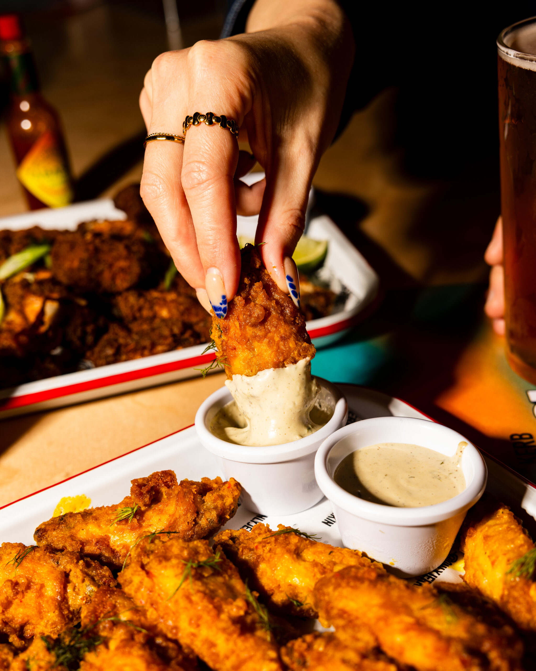 A hand with painted nails dips a crispy fried chicken wing into creamy sauce at a lively Sports Bar in Shoreditch, with more wings and a cold glass of beer nearby on the table.