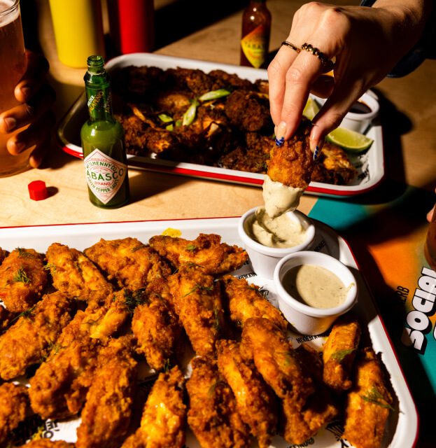 Close-up of trays filled with crispy fried chicken wings at a lively Sports Bar in Shoreditch. One hand dips a wing into sauce, another holds a drink. Bottles of hot sauce and condiments are scattered on the table.