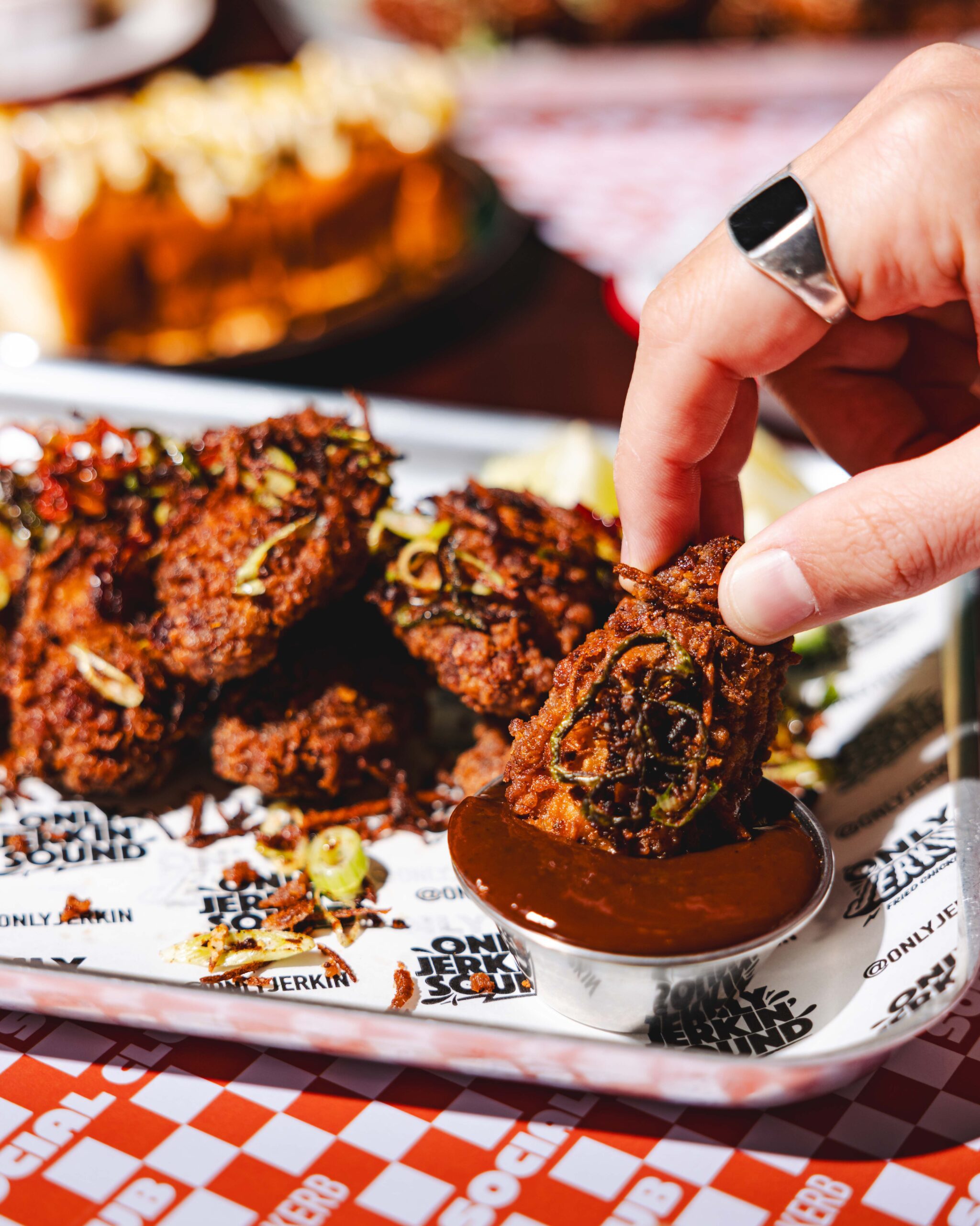 A hand dips a crispy fried chicken wing into a cup of dark sauce on a tray lined with branded paper, with more wings and a blurred dish in the background.