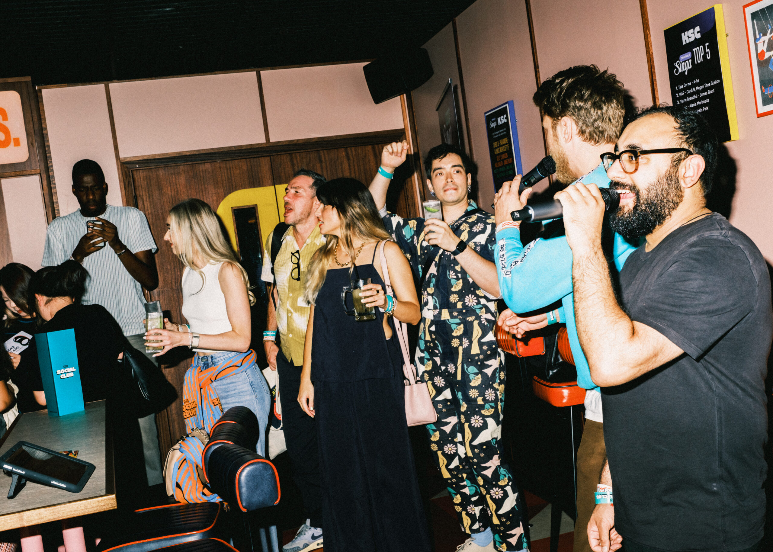 A group of people stand and cheer in a lively, colourful bar. Some hold drinks, one man sings into a microphone, and others are smiling, waving book darts as they interact and celebrate, creating a festive, energetic atmosphere.