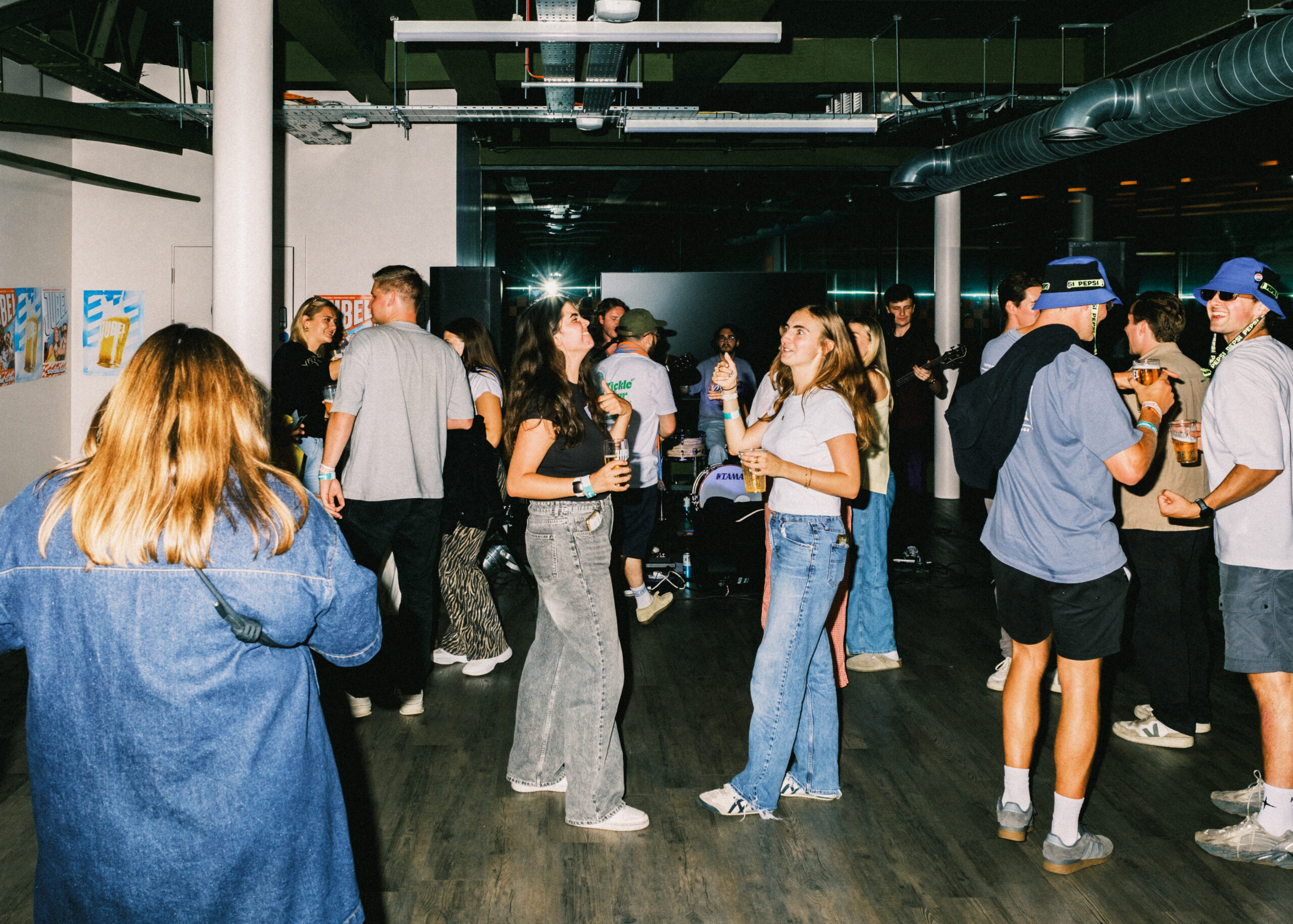 A group of casually dressed young adults socialise, drink, and dance in a modern indoor space with exposed pipes, art on the walls, and book darts scattered on tables. The lighting is bright and some people are looking towards the camera.