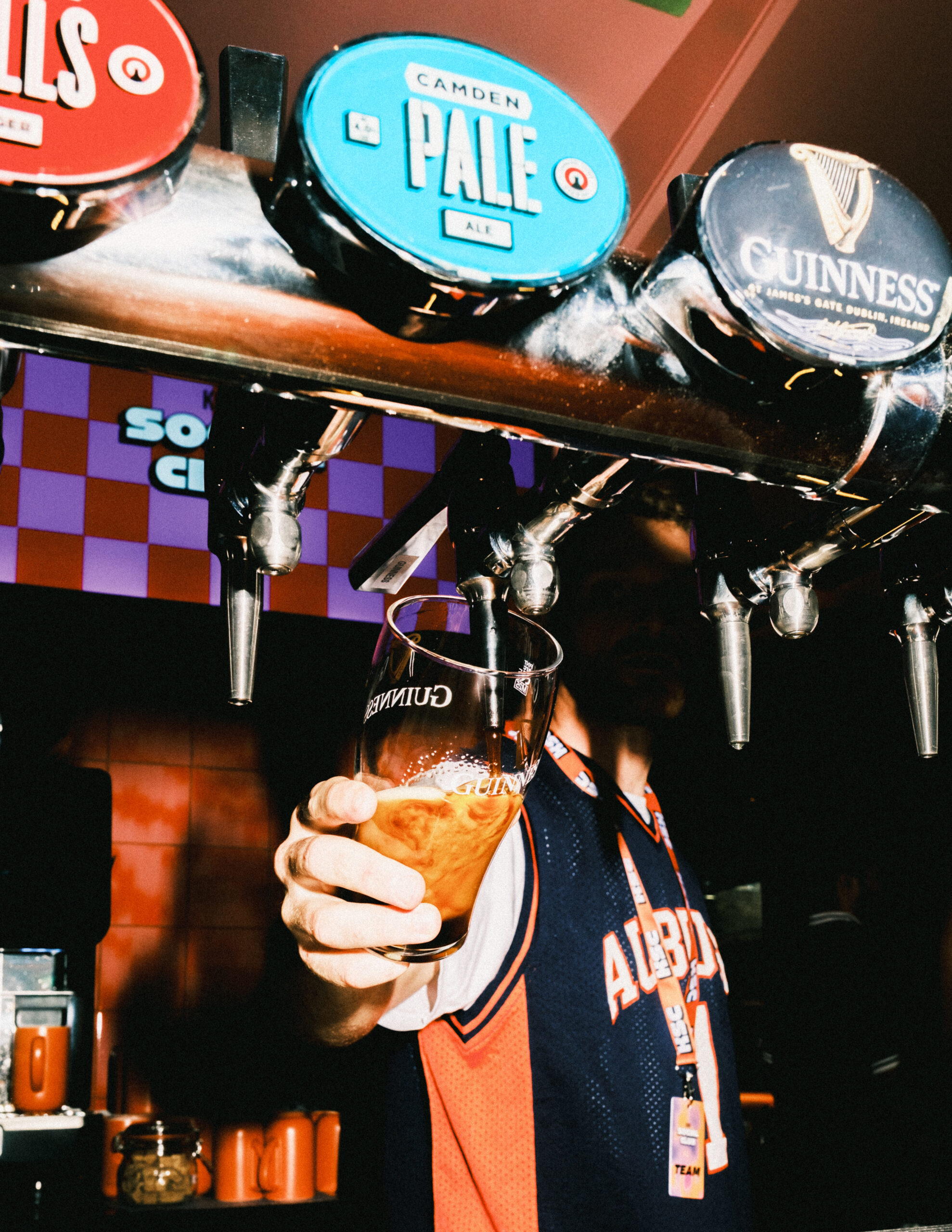 A person holds a glass under a Guinness beer tap, filling it with beer at a pub. Other taps labelled Camden Pale and Guinness are visible above, creating a lively setting perfect for celebrating the excitement of World Cup 2026.
