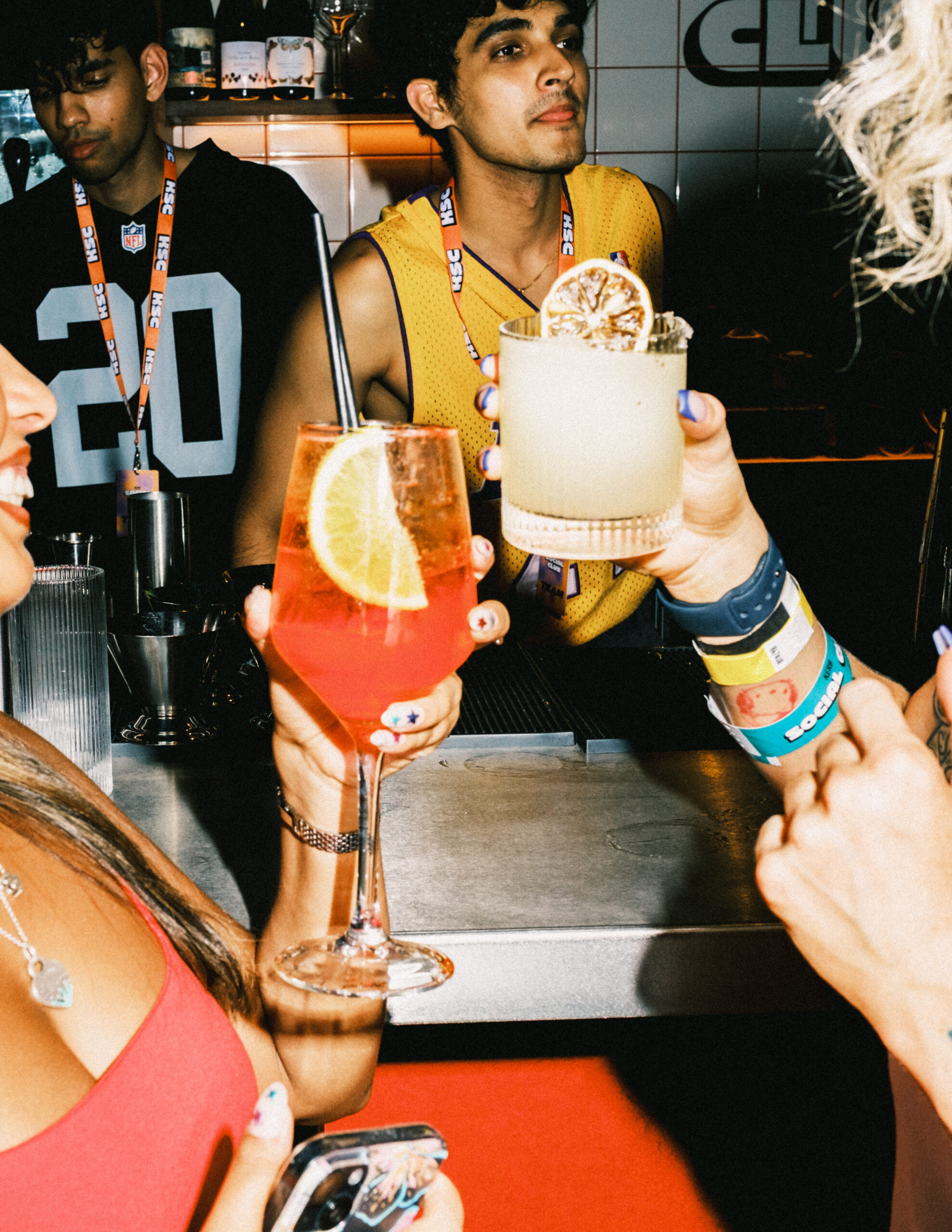 Two people clink colourful cocktails in a lively bar, one with a red drink garnished with an orange slice and the other with a pale drink and dried citrus. A barman in a yellow jersey, ready for World Cup 2026 fever, watches from behind the counter.