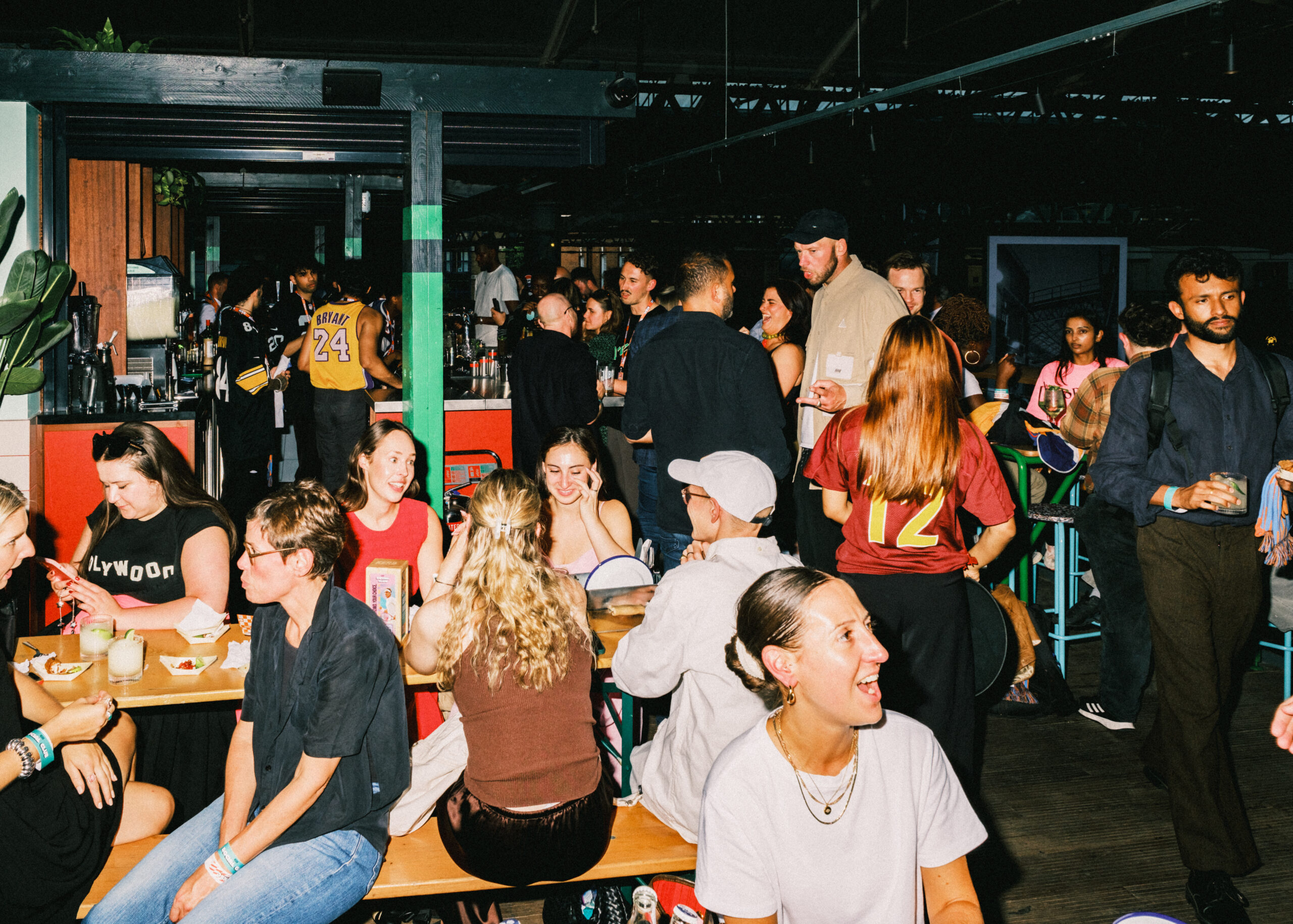 A lively indoor gathering with people socialising, eating, and drinking around tables and a bar. Some wear sports jerseys, while others show off book darts as conversation starters. The atmosphere is casual and energetic under bright lighting.