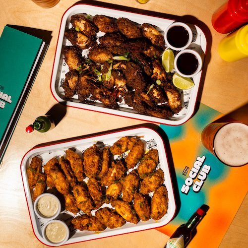 Two trays of chicken wings—one with a dark, crispy coating and lime wedges, the other golden with dipping sauce. Drinks, condiments, and Social Club menus sit on the wooden table, ready for a perfect World Cup 2026 viewing party.