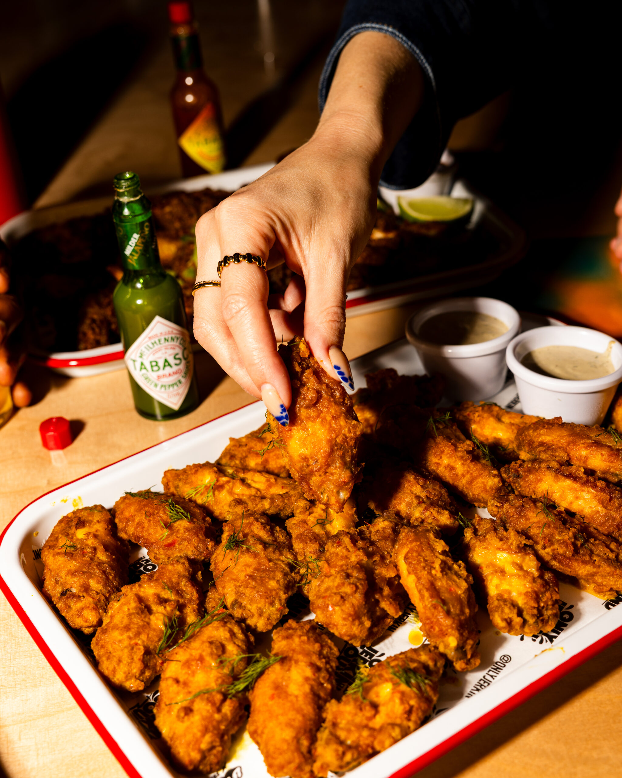 A hand reaches for a crispy fried chicken wing from a tray filled with wings, garnished with herbs. Nearby are two dipping sauces, a bottle of Tabasco sauce, and another tray of wings in the background.