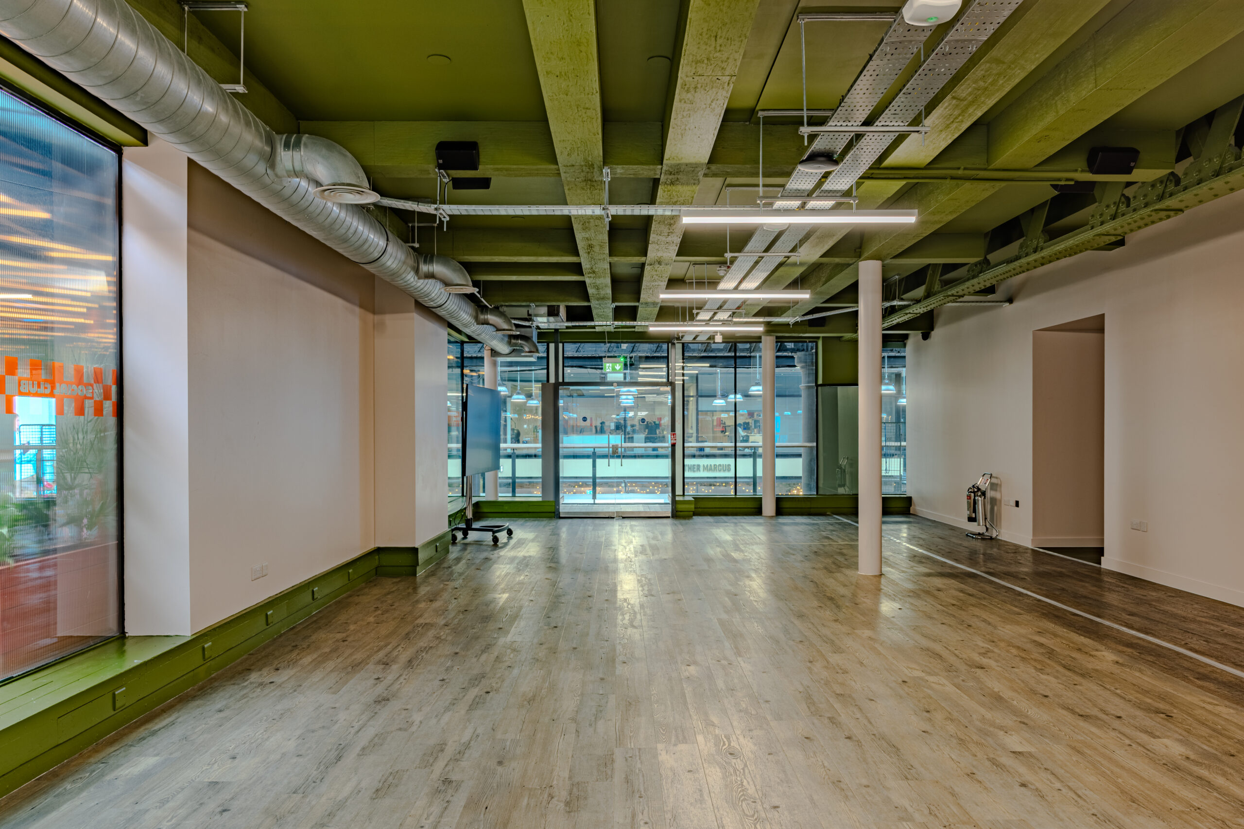 A modern, empty office space with wooden flooring, large windows, exposed ceiling pipes, and green-painted ceiling beams. Bright lighting and a glass door create a crisp atmosphere—perfect for creative minds or arranging book darts in style.