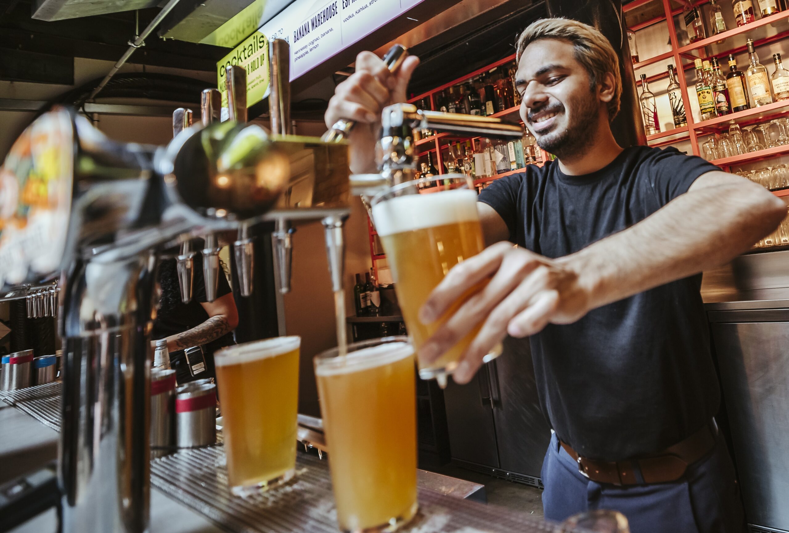 A barman with light brown hair pours draught beer into a glass behind a bar, with two full glasses of beer on the counter and shelves of spirits bottles in the background, ready for fans to cheer during the World Cup 2026.