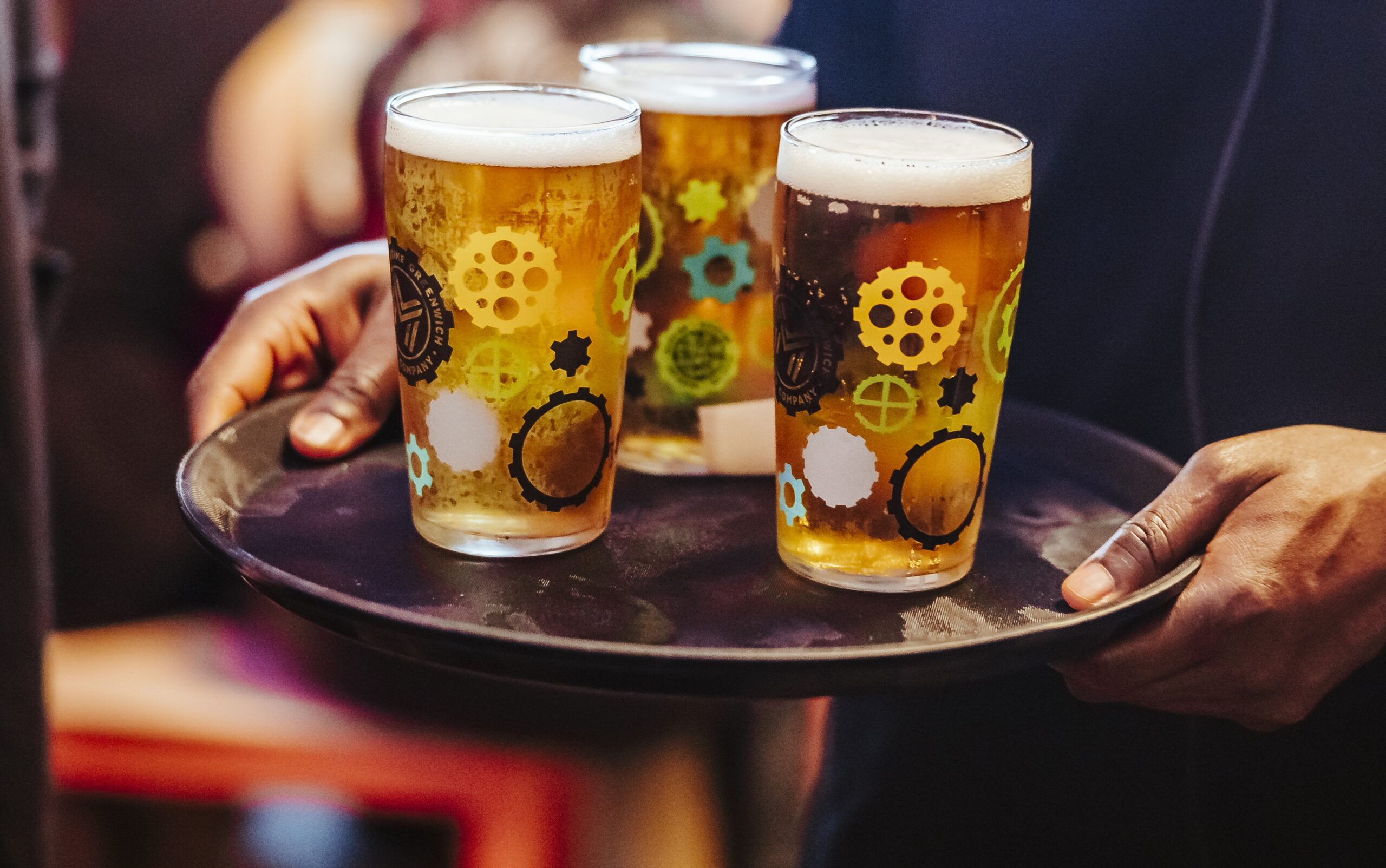 A person holds a tray with three pint glasses of beer, each glass decorated with colourful gear and circle patterns, as they celebrate in a lively indoor setting during the build-up to World Cup 2026.