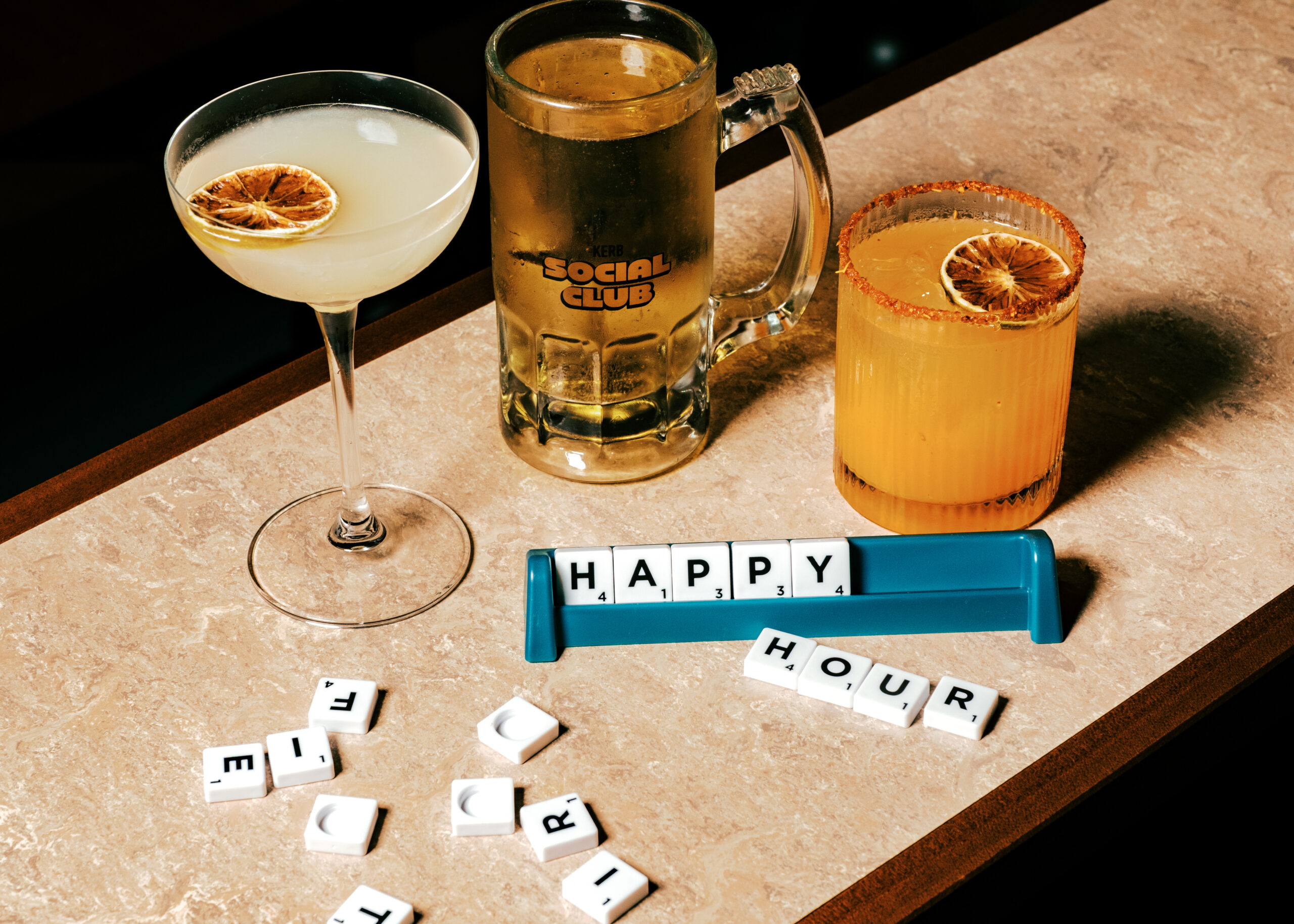 Three drinks on a bar—one cocktail with dried lime, one beer tankard labelled THE SOCIAL CLUB, and an orange cocktail—sit beside Scrabble tiles spelling HAPPY HOUR. Scattered tiles decorate the surface, inviting you to Film Club KERB.