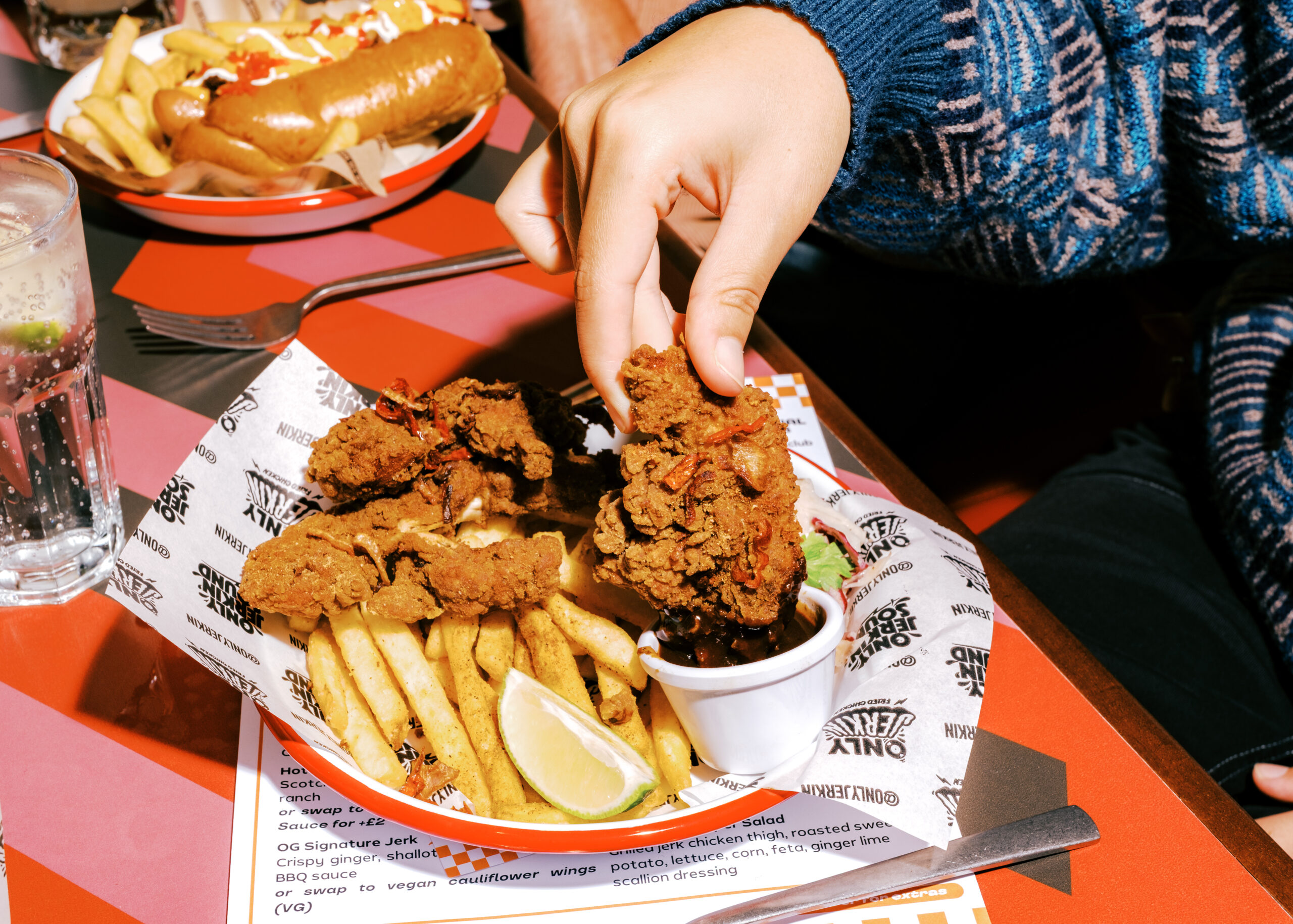 A person dips a piece of fried chicken into sauce, with more fried chicken, chips, and a lime wedge on a plate. There is a glass of water and another plate of food in the background.