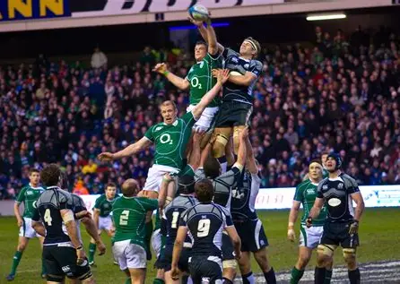Rugby players from two teams, one in green and one in dark blue, jump and compete for the ball during a Six Nations line-out, with a large crowd watching from the stadium stands near the buzzing KERB food stalls.