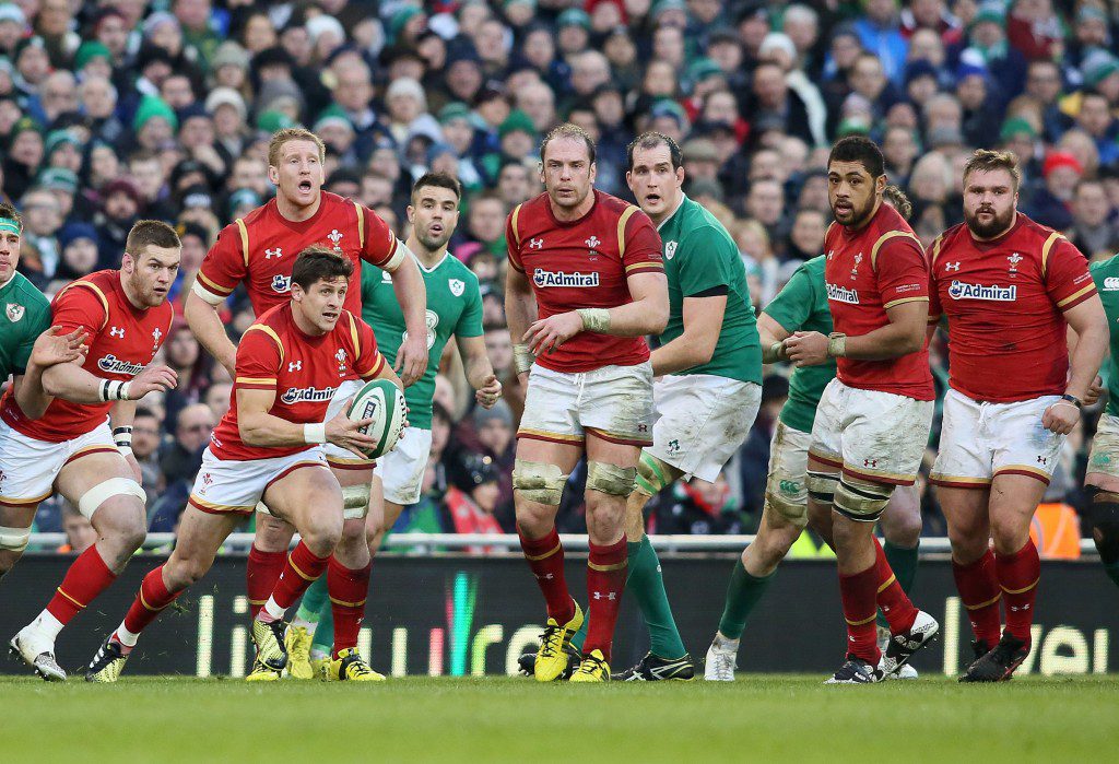 A group of rugby players in red jerseys face opponents in green during a Six Nations KERB match, with one red team player running whilst holding the ball. The crowd is visible in the stadium background.
