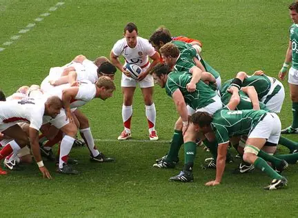 Two rugby teams, one in white and one in green, face each other in a scrum on a grassy pitch during a thrilling Six Nations match, as the referee stands ready to throw the ball in between them.