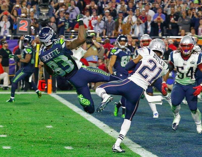 A Seattle Seahawks player attempts to catch a pass in the end zone whilst a New England Patriots defender blocks him during a Super Bowl 2026 match-up, with spectators in the background.