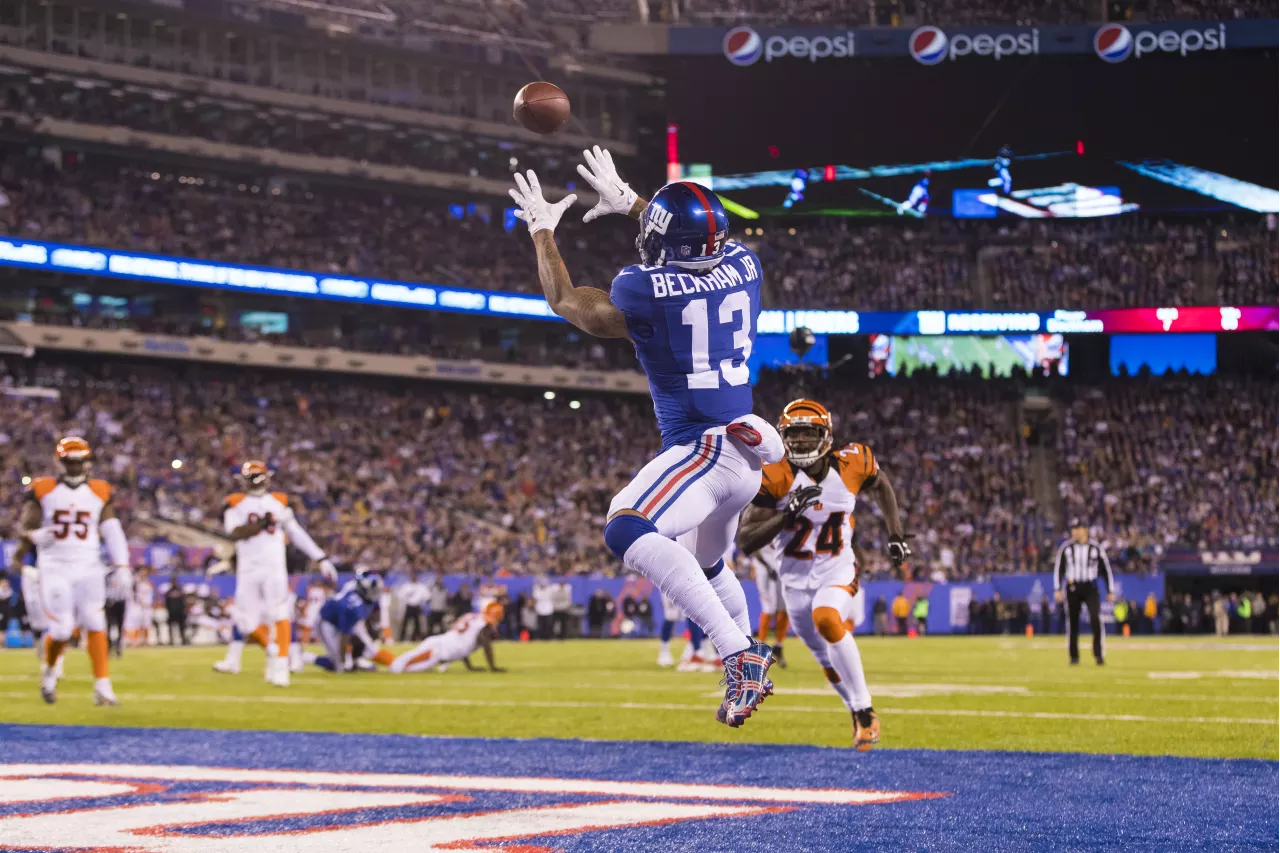 A footballer in a blue shirt leaps to catch a pass in the end zone during the 2026 Super Bowl, while a defender in white approaches. The stadium is packed with fans and bright lights illuminate the pitch.