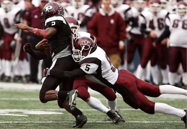 A footballer in a black kit is being tackled by an opposing player in a white and maroon kit during a match, capturing the intensity seen on the road to Super Bowl 2026, with other players and coaches visible in the background.