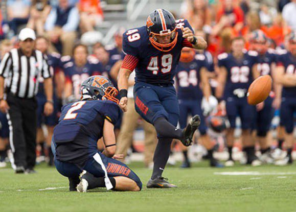 A footballer in a blue kit, number 49, is kicking a football held by teammate number 2 during a match—perhaps dreaming of making plays like this in the 2026 Super Bowl. Other players and an official are visible in the blurred background.