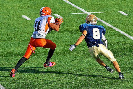 A footballer in an orange and white kit catches a pass while being pursued by a defender in blue and gold, showcasing the intensity supporters can expect at Super Bowl 2026 on the vibrant green pitch.