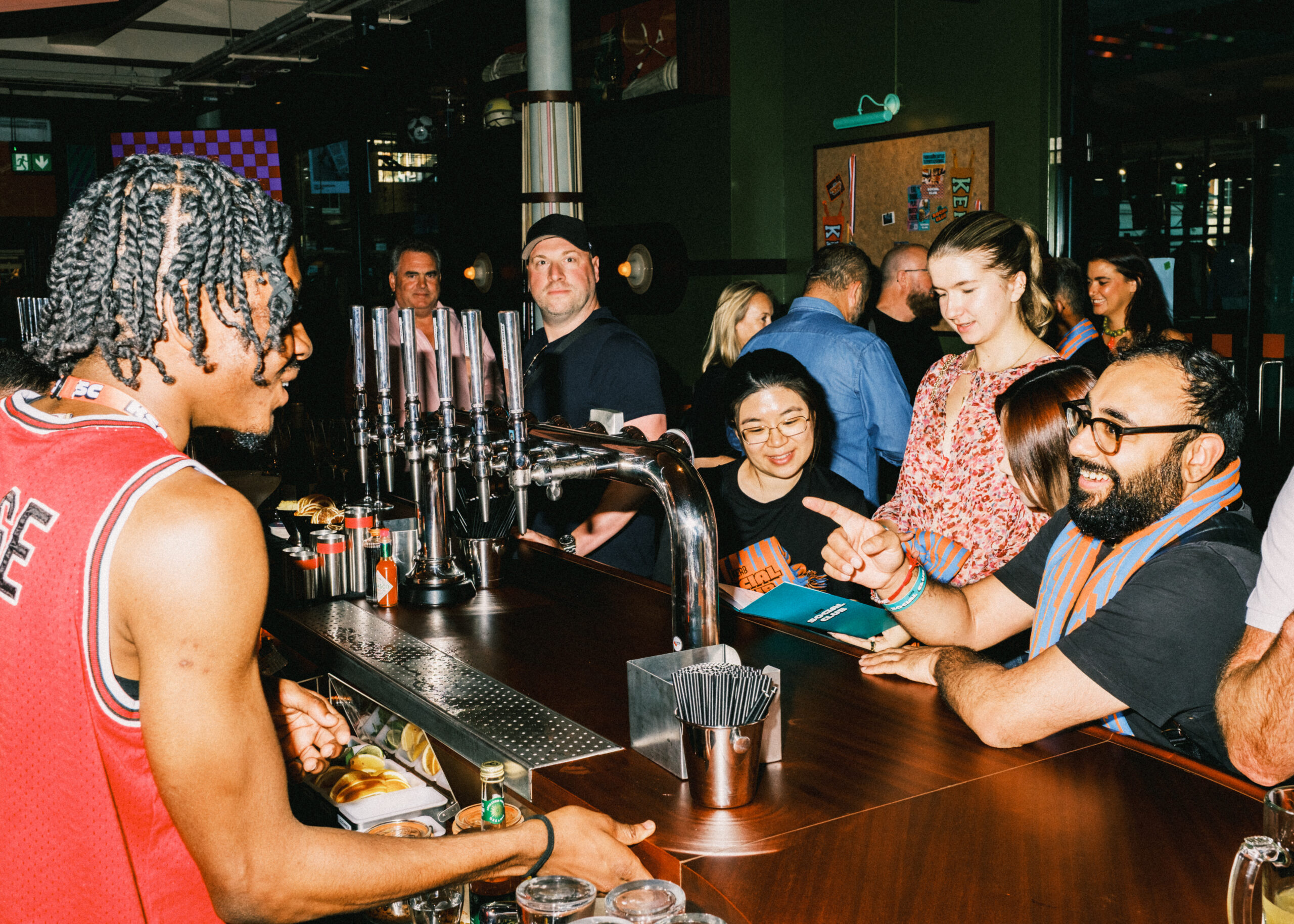 A barman in a red jersey serves drinks to a lively group of people at a bar; several customers smile, chat, and gesture as they await the Super Bowl 2026 action at the counter.