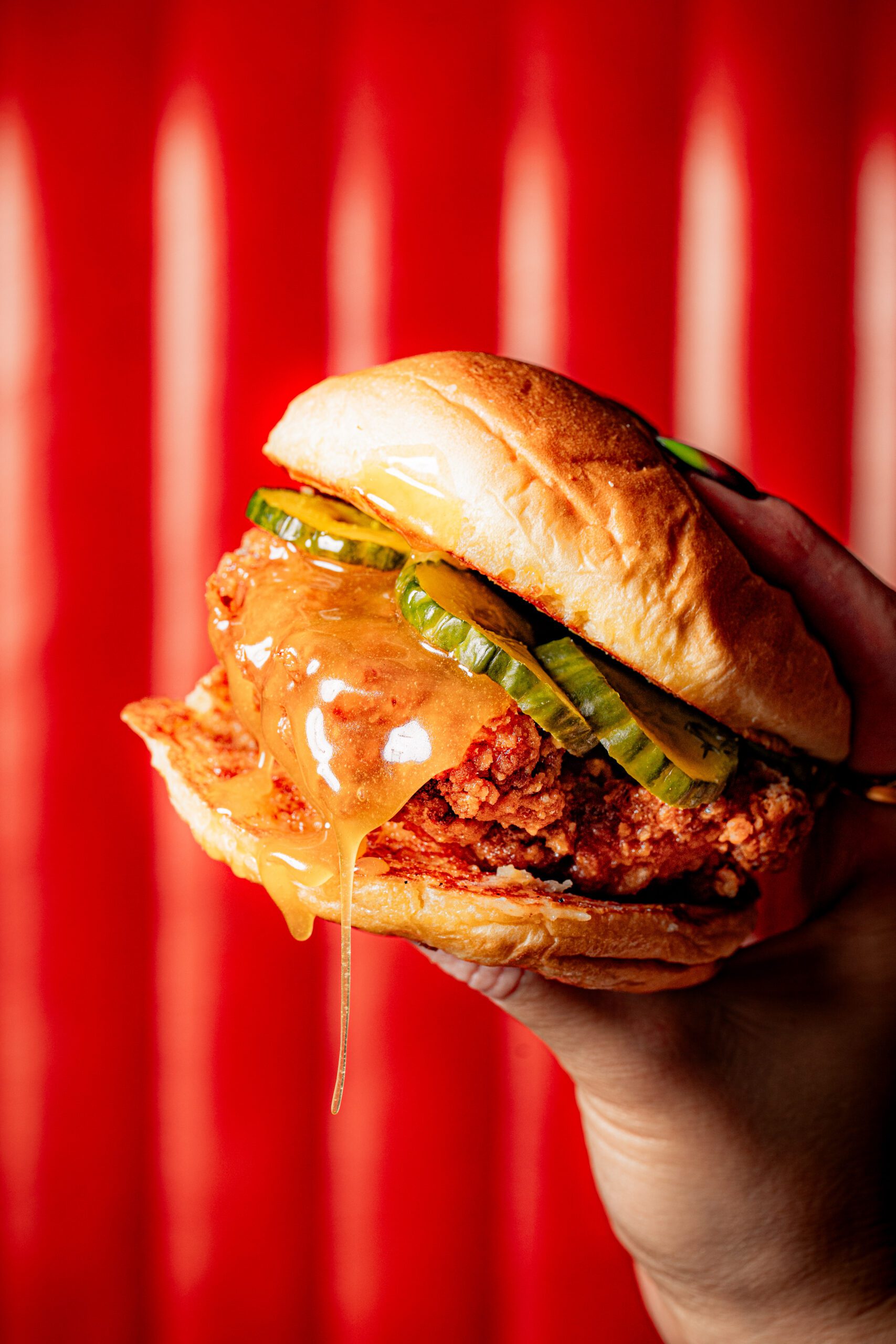 A hand holds a crispy fried chicken sandwich with pickles and melted cheese dripping down the side, set against a vibrant red background—highlighting OJ's NEW MENU.