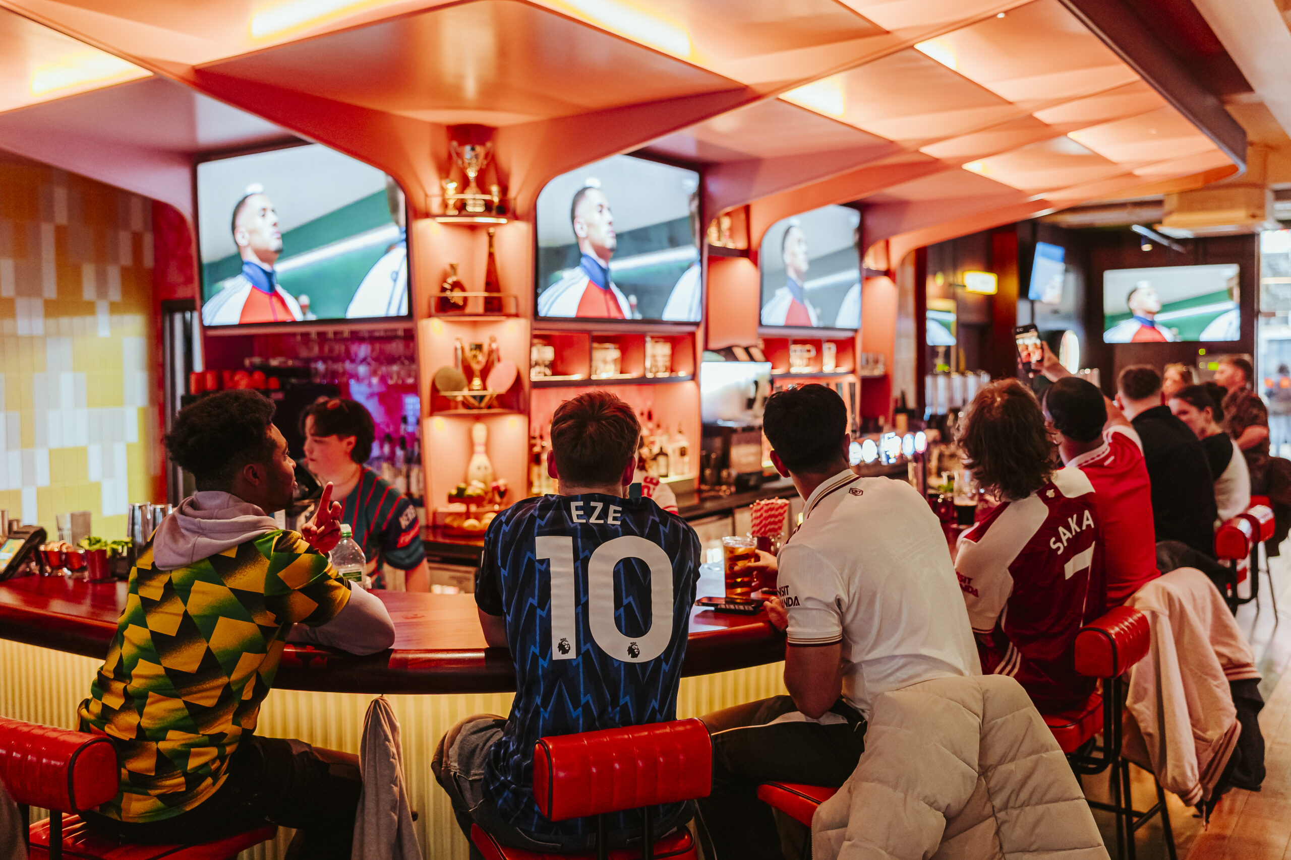 A group of people sit at a bar with drinks, watching a World Cup 2026 football match on several large TVs. Some wear football shirts and appear engaged in the game, as bar shelves behind display trophies and sports memorabilia.
