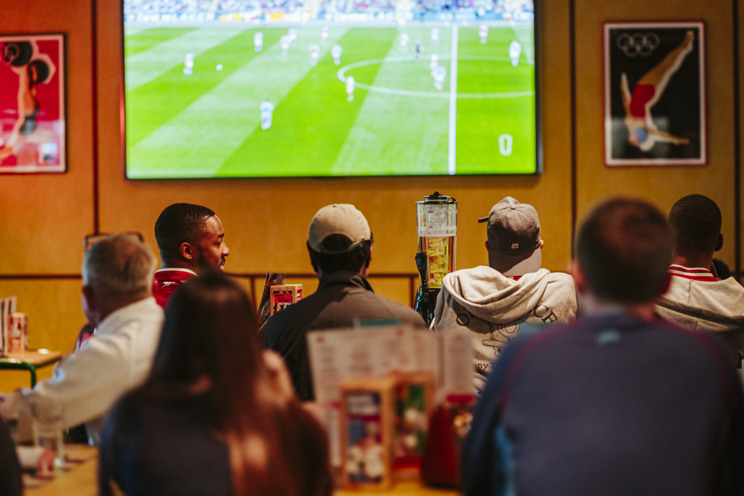 A group of people sit at tables in a bar or restaurant, watching a World Cup 2026 football match on a large TV screen mounted on the wall. Menus and drinks are visible on the tables in the foreground.