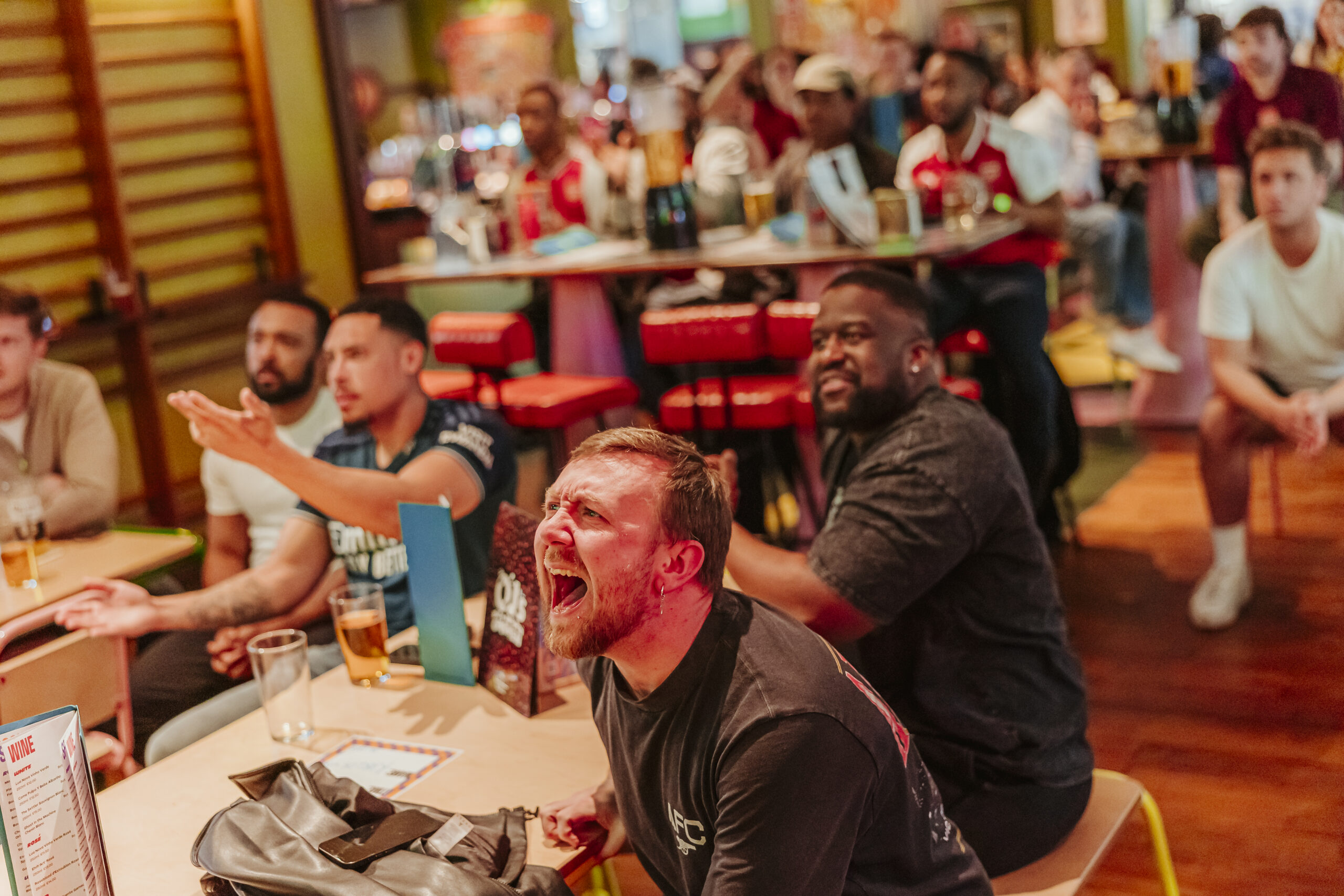 A group of men sits at tables in a lively pub, watching something off-screen—likely the World Cup 2026. One man in the foreground shouts passionately, while others gesture or react with intense focus. Drinks and menus are on the tables.