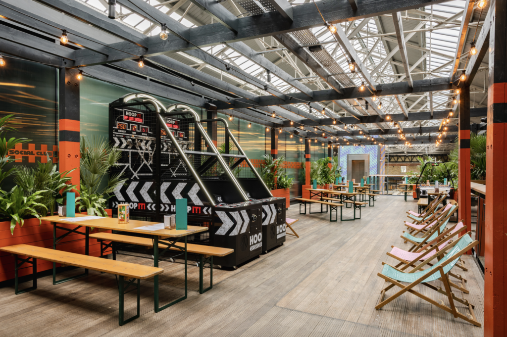 A bright indoor leisure space with wooden tables, benches, deckchairs, book darts for avid readers, potted plants, fairy lights, and a basketball arcade game under a glass roof.