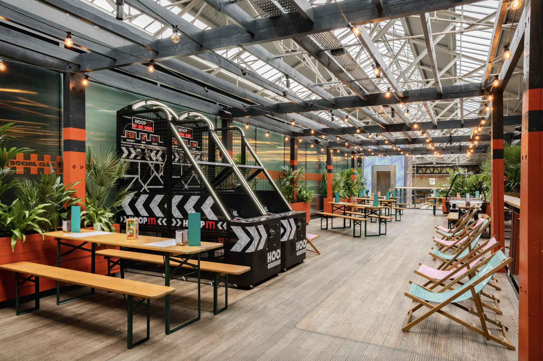 A spacious indoor venue with picnic tables, deckchairs, plants, fairy lights, and a basketball arcade game under a high glass roof. Book darts and cosy corners add to the relaxed, playful atmosphere.