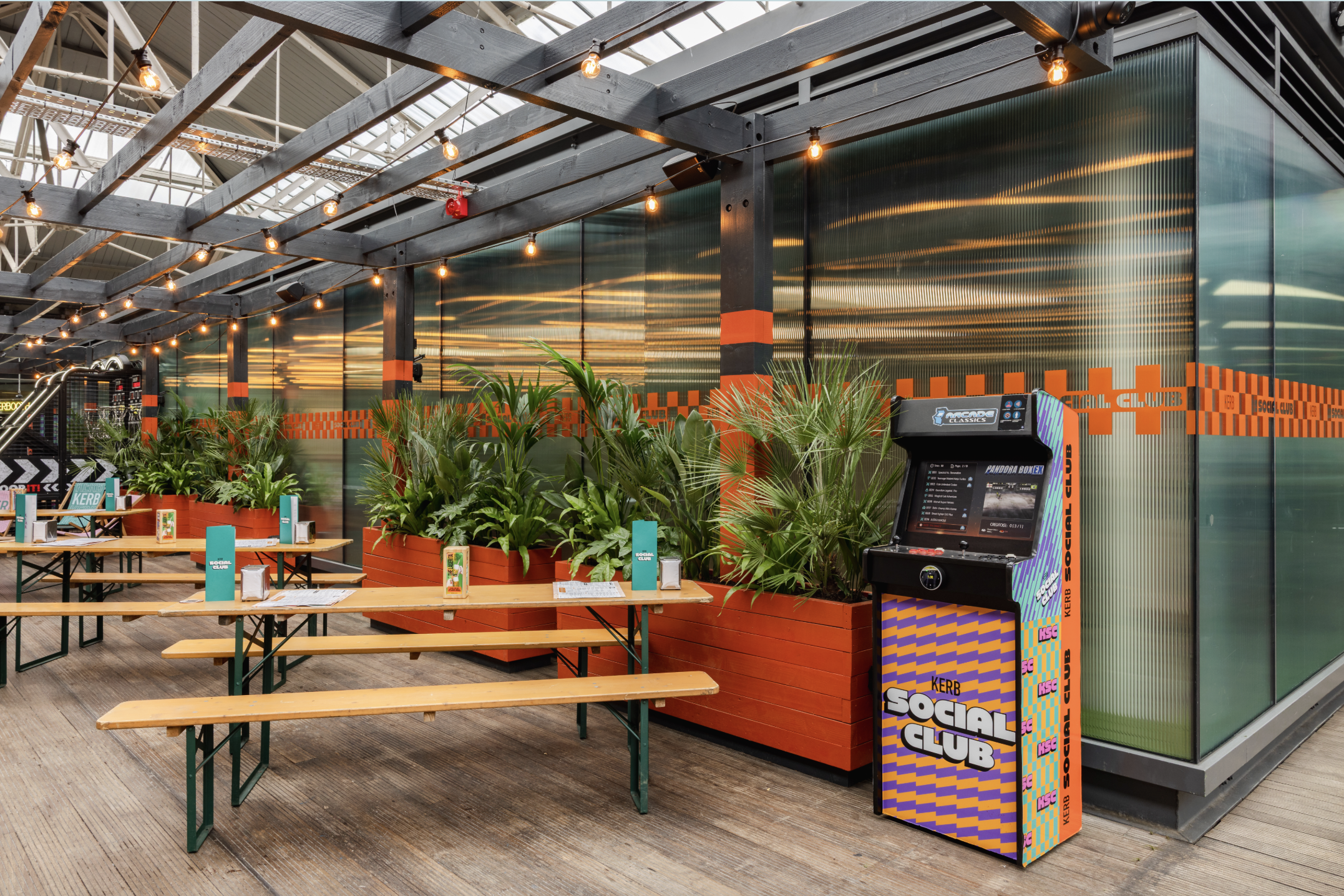 A modern indoor space with long wooden tables and benches, potted green plants, fairy lights, and a colourful arcade machine labelled Social Club. Book darts rest on the tables beneath a glass wall with orange accents.