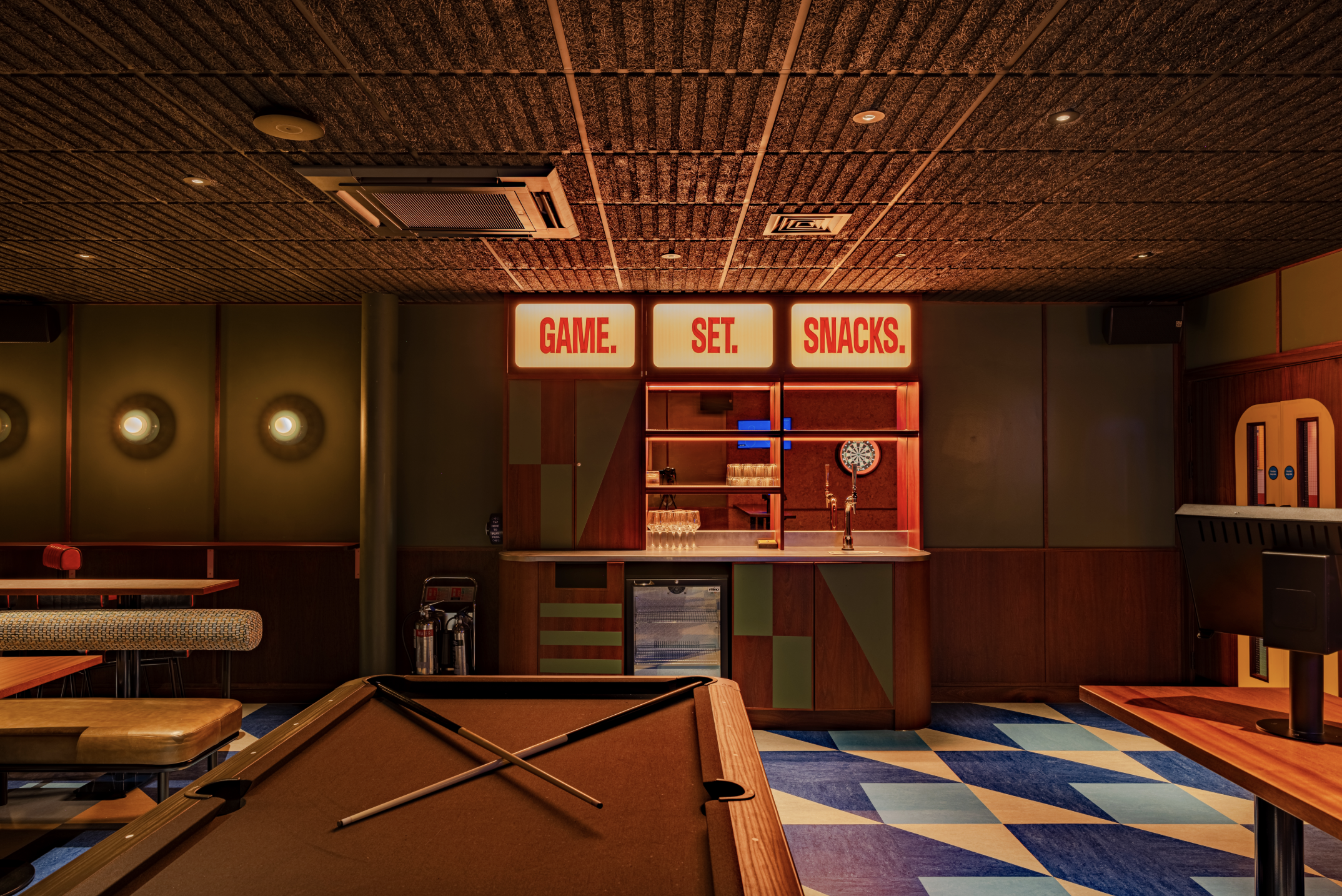 A retro-style games room with a pool table, crossed cues, book darts displayed by the snack bar sign GAME. SET. SNACKS., round wall lights, and blue geometric-patterned flooring.