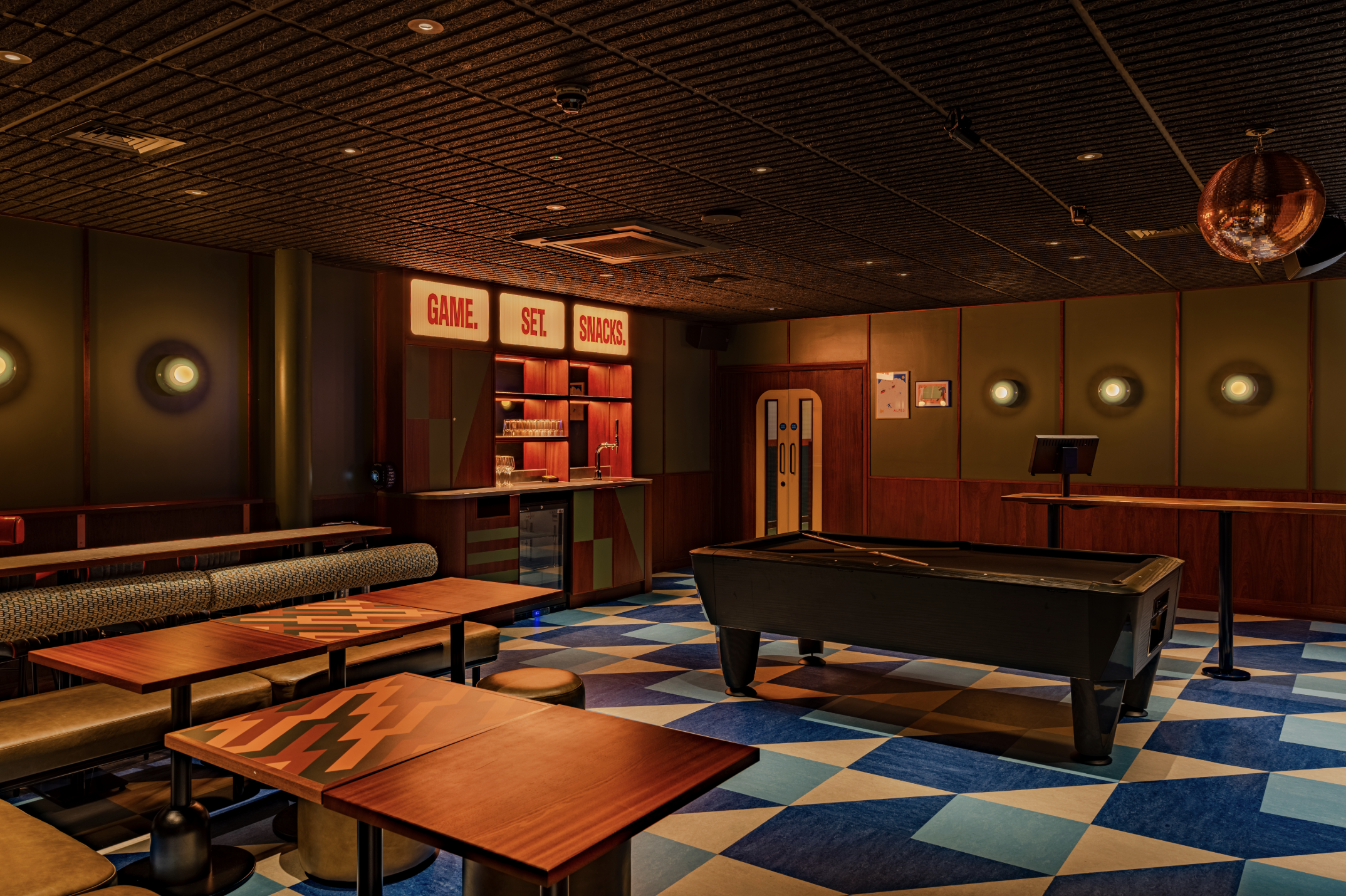 A stylish games room features a pool table, geometric blue and white floor tiles, wooden tables, cosy seating, wall lights, book darts by the snack bar with a “GAME SET SNACK” sign. The room feels modern and inviting.