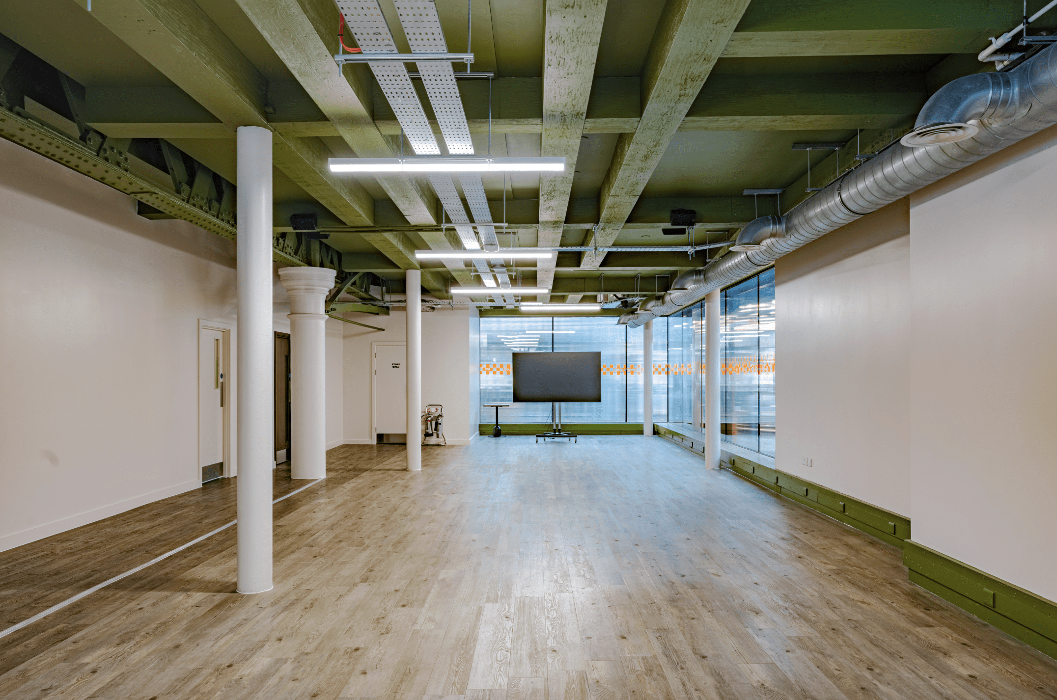 An empty modern office space with wooden floors, green-painted exposed ceiling beams, and white walls. Large windows and a glass partition are at the far end; a TV screen on a stand is placed near book darts scattered by the windows.