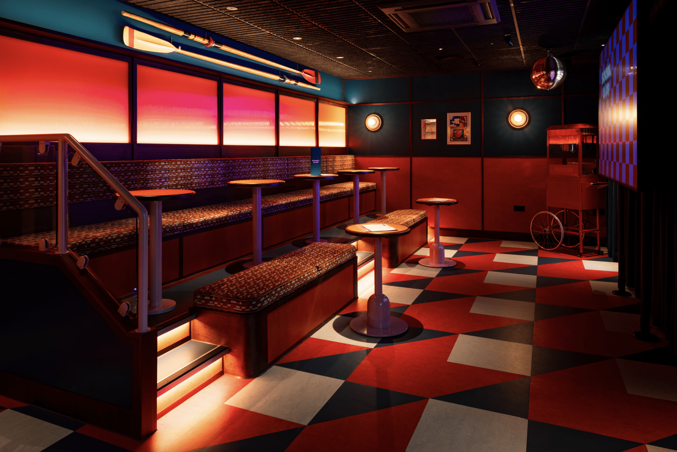 A retro-style lounge with patterned benches, small tables, red and blue geometric flooring, vibrant lighting, framed posters on the wall, a popcorn trolley, book darts as quirky accents, and a disco ball hanging from the ceiling.