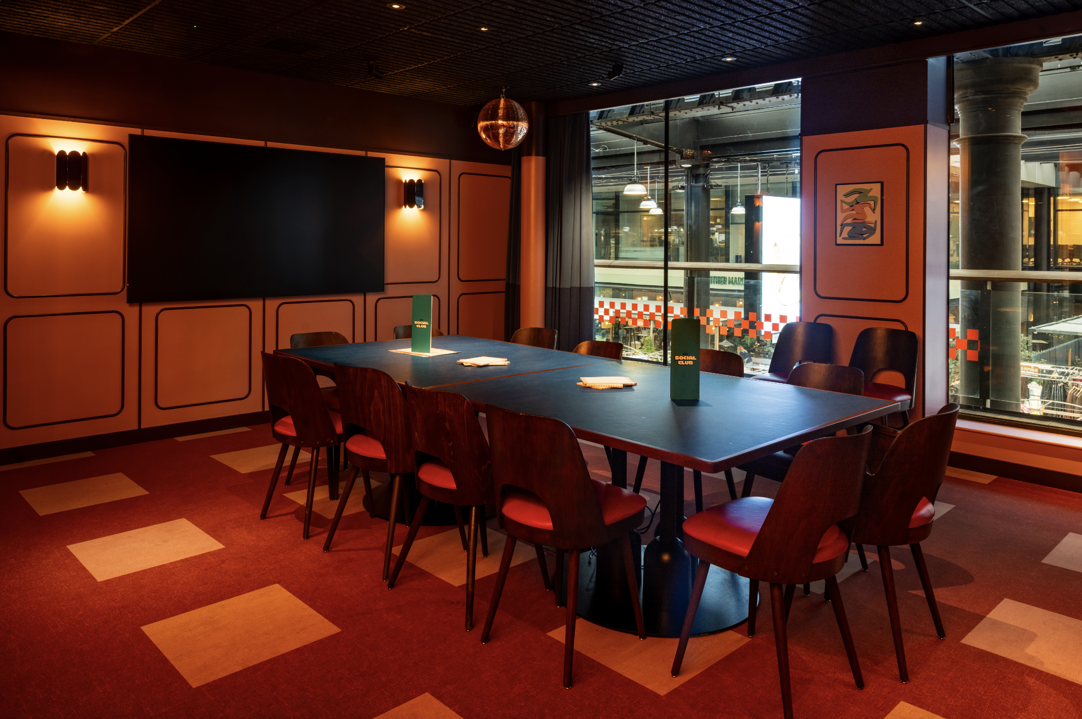 A modern meeting room with a long black table surrounded by red-cushioned chairs, dim wall lights, a large screen, and floor-to-ceiling windows overlooking an industrial space. Papers, folders, and book darts are scattered on the table.