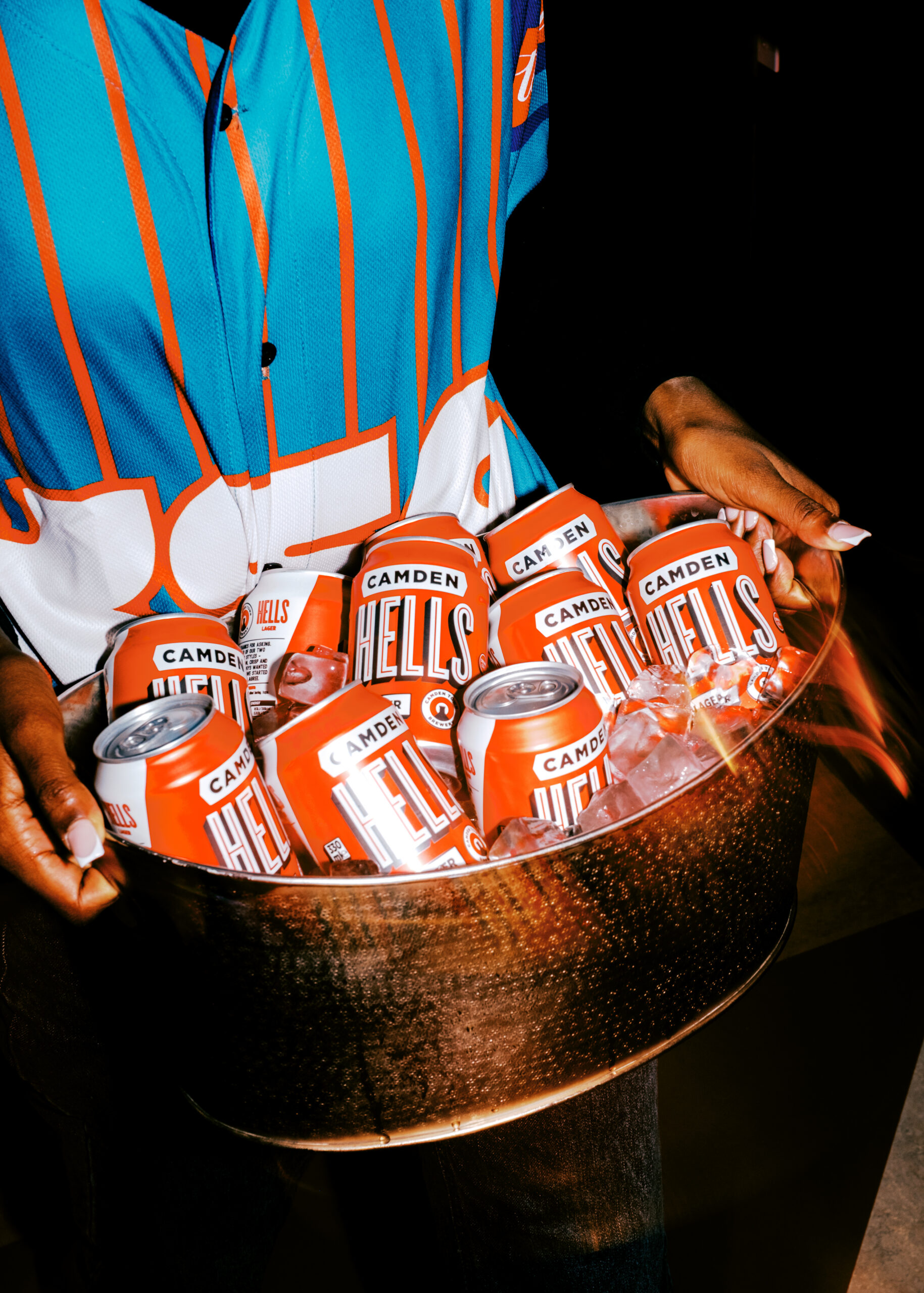 A person in a blue sports jersey holds a metal tub filled with ice and several cans of Camden Hells beer, ready to celebrate the excitement of World Cup 2026 against a dark background.