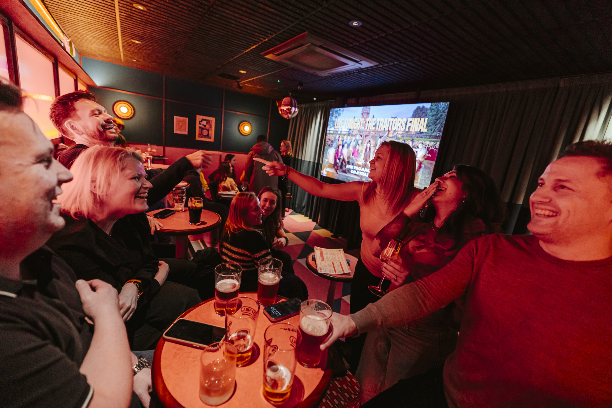 A group of people laugh and celebrate together at a pub, raising drinks and clinking glasses whilst watching a sports event on a large screen in a cosy, warmly lit space—perfect for those who want to book a private room for their celebration.